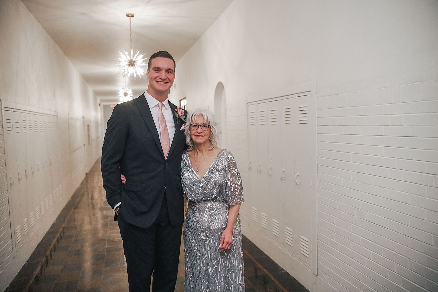 Two people stand together in a white hallway with lockers, dressed in formal attire for a special occasion.