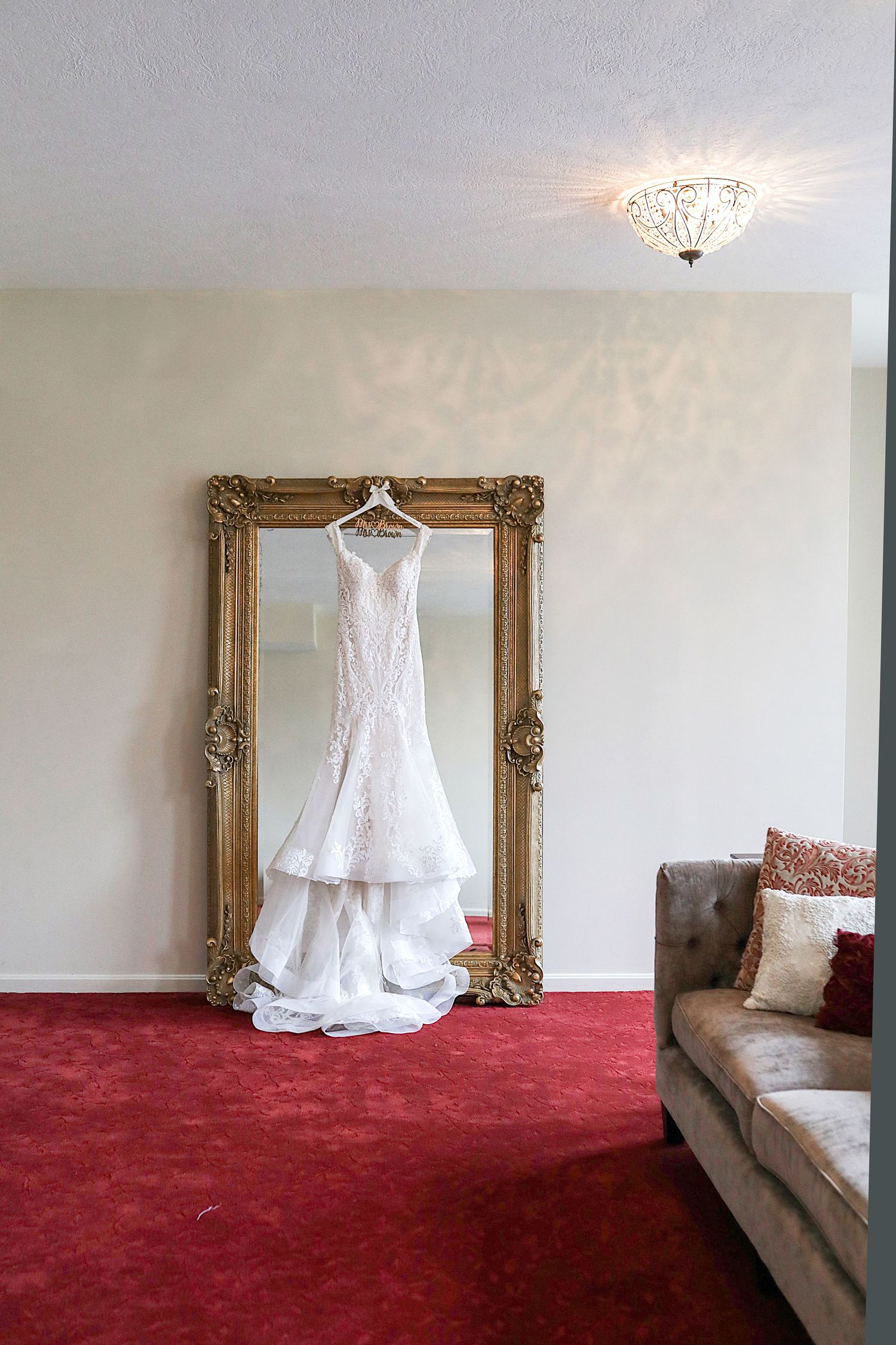 A white wedding dress hangs on an ornate gold mirror frame in a room with red carpet and a gray couch.