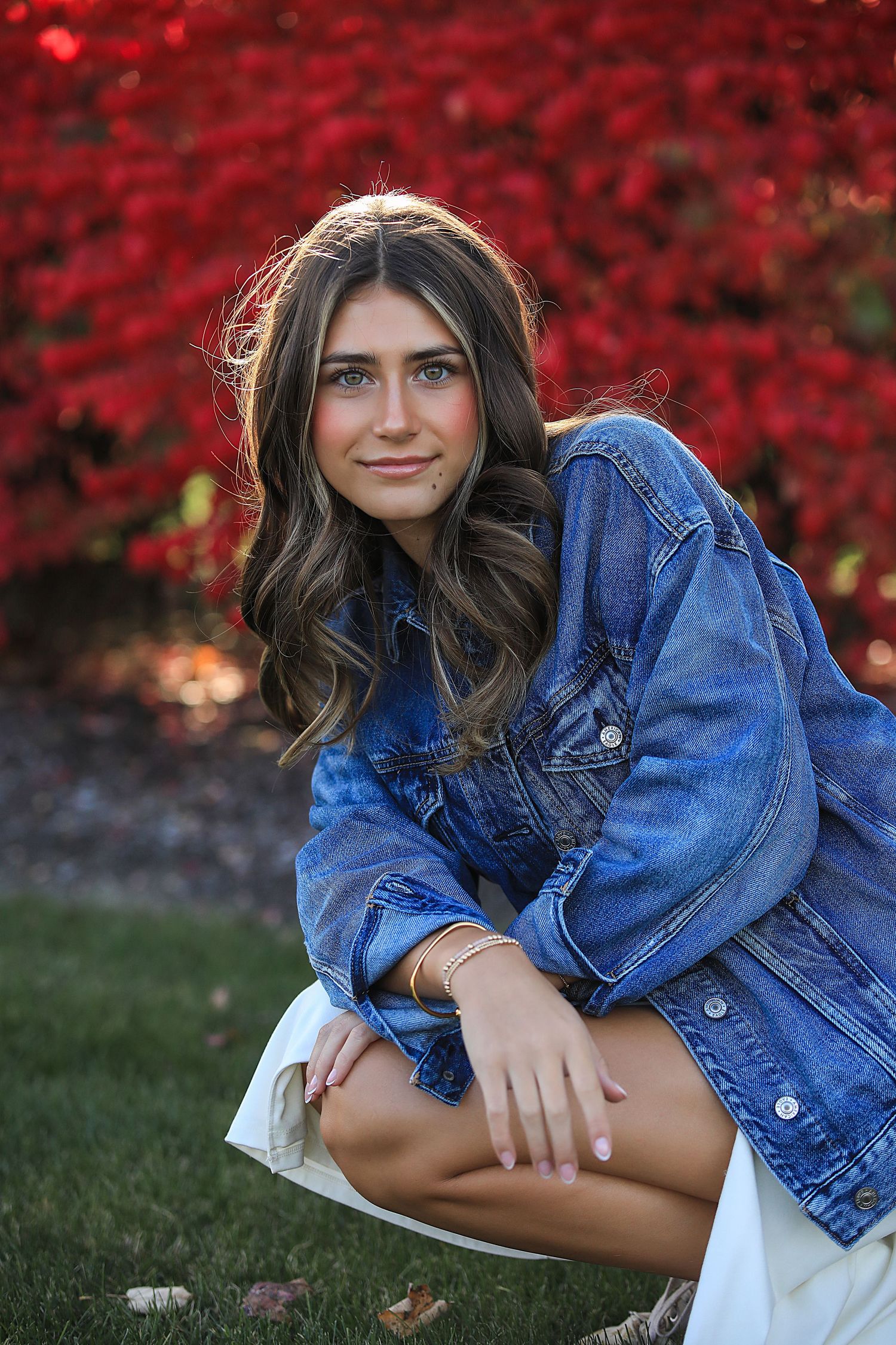 Portrait of someone in a denim jacket crouching on grass with vibrant red autumn foliage in the background.