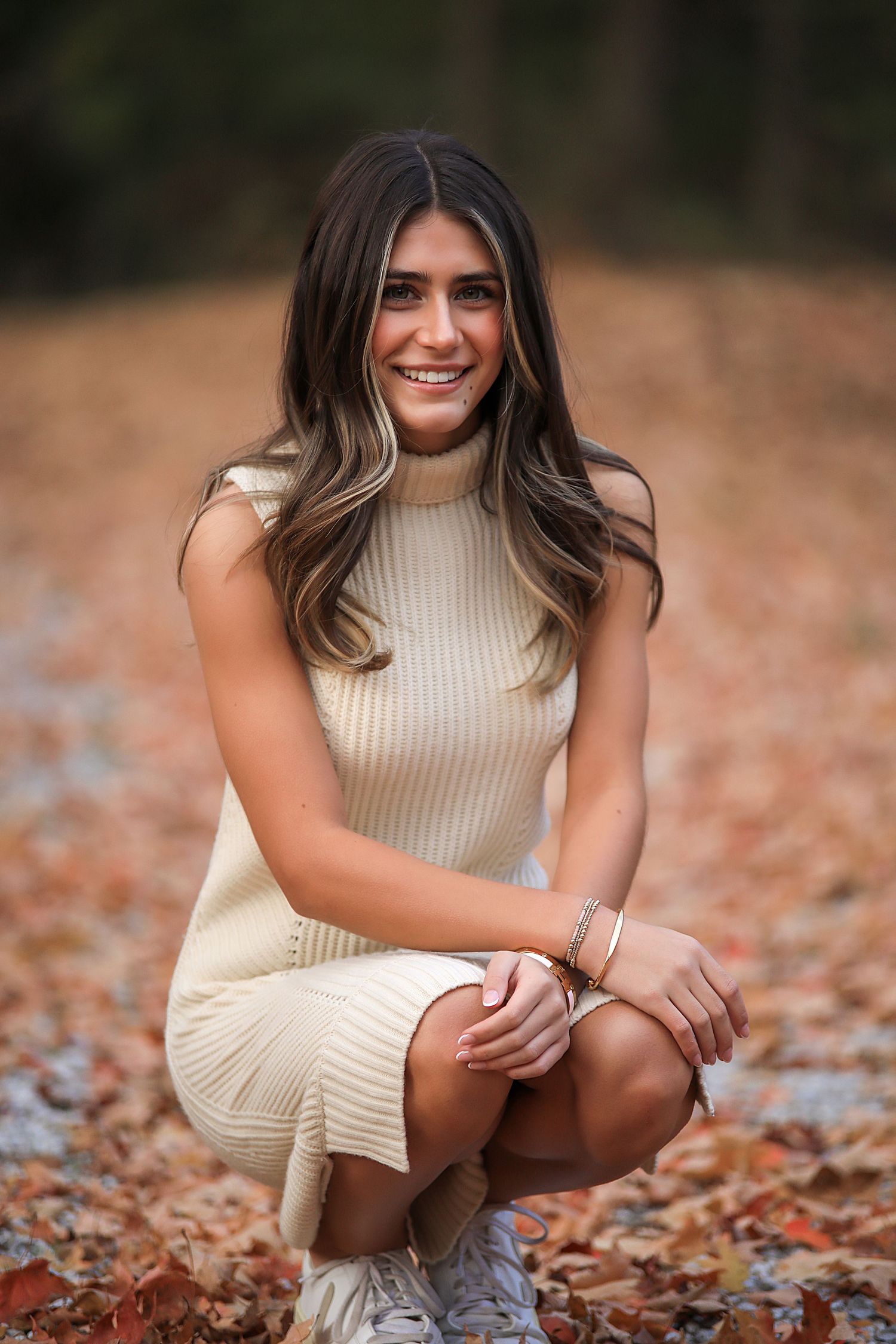 Young woman wearing cream turtleneck sweater sits on autumn leaves in outdoor portrait series.
