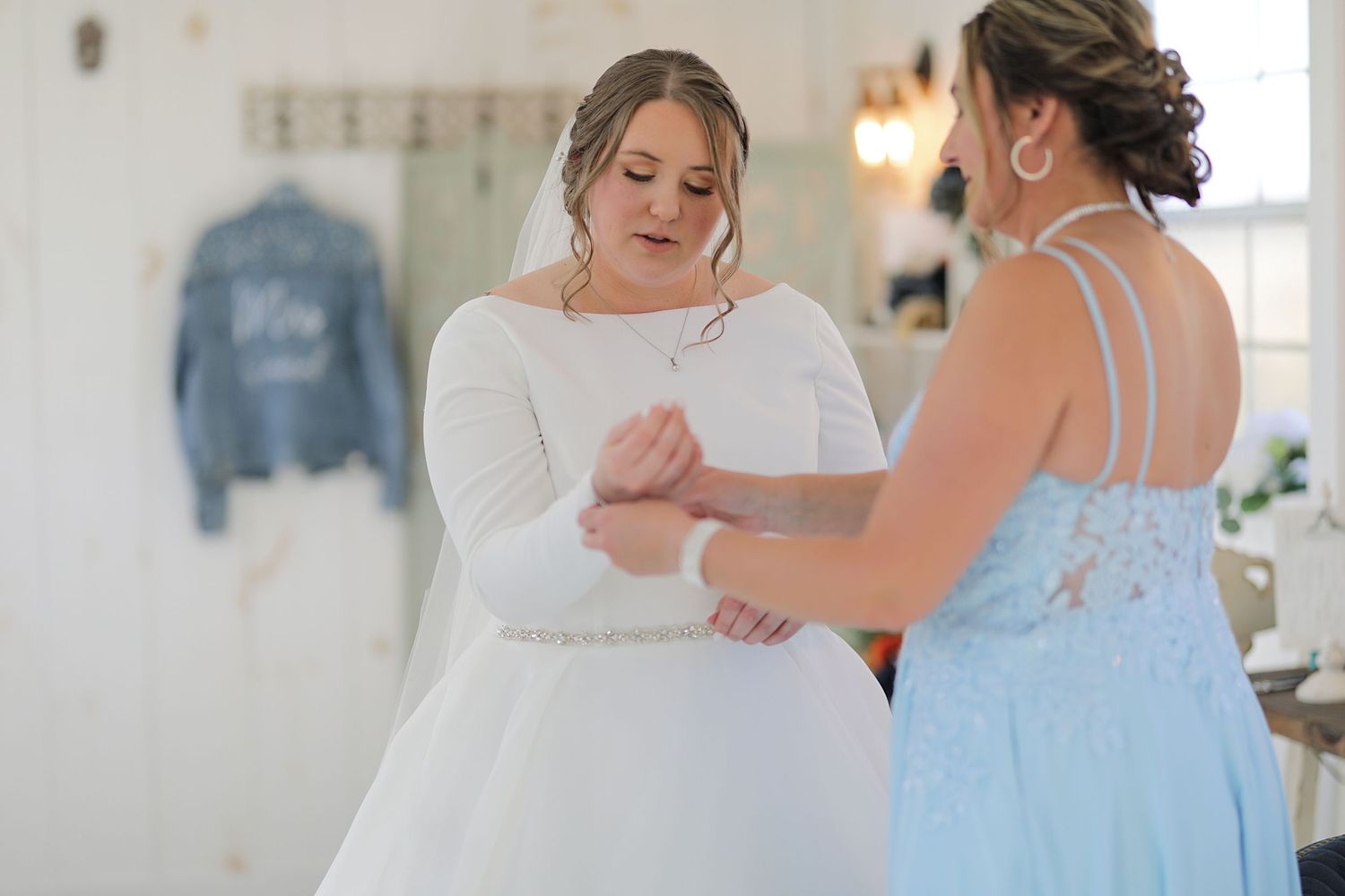 Two people in wedding attire getting ready in a soft-lit dressing room with a blue denim jacket visible in background.