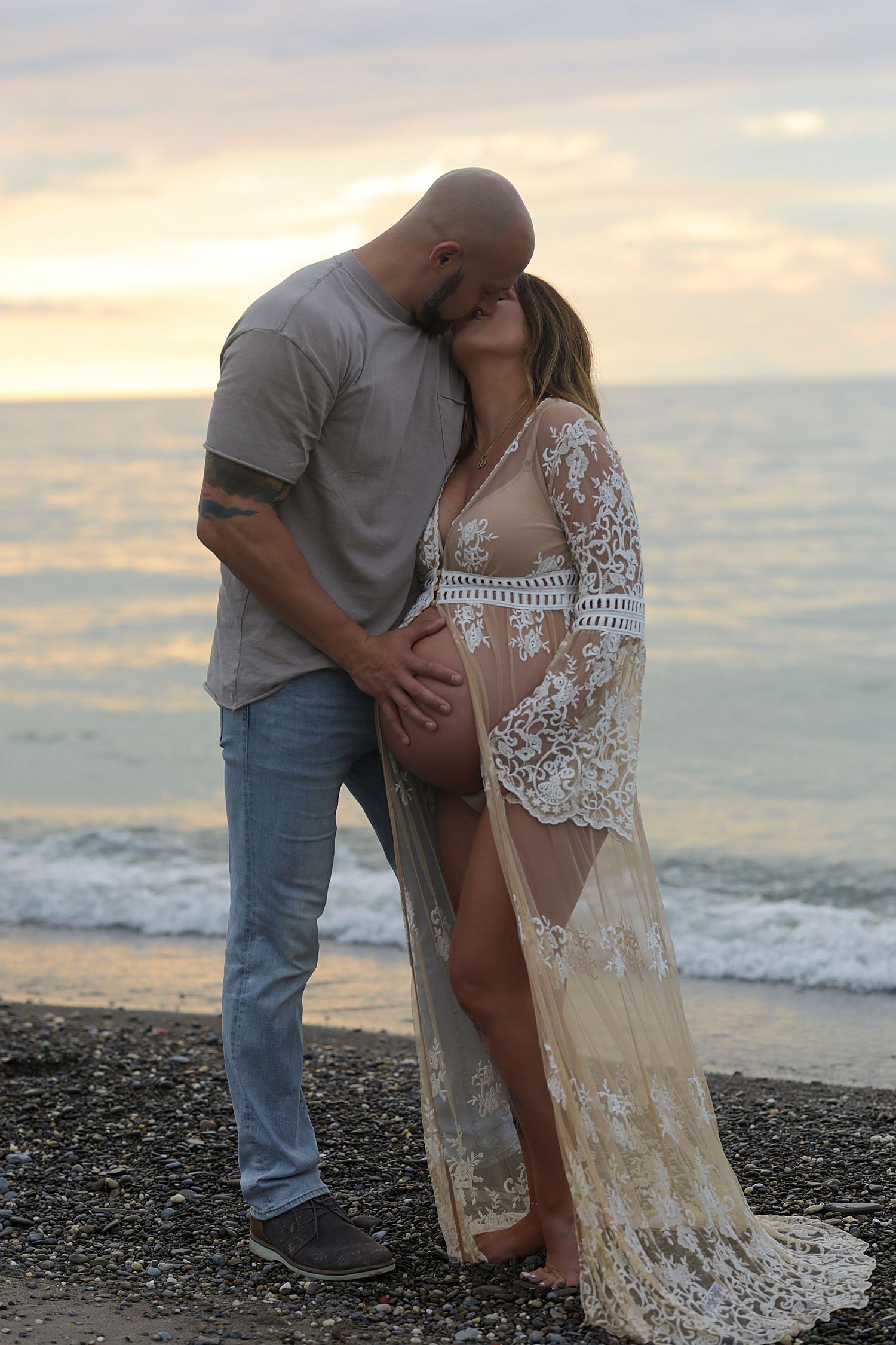 A couple shares an intimate moment at sunset on a beach, with the woman wearing a long lace robe and the man in casual attire.