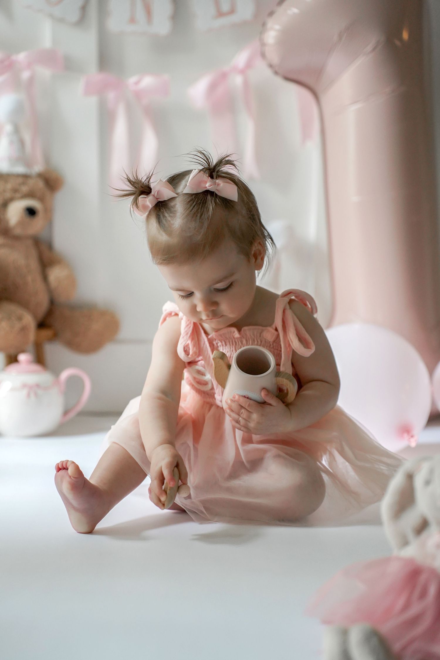 Sweet baby in pink dress plays on soft white backdrop with stuffed animal in dreamy nursery setting.