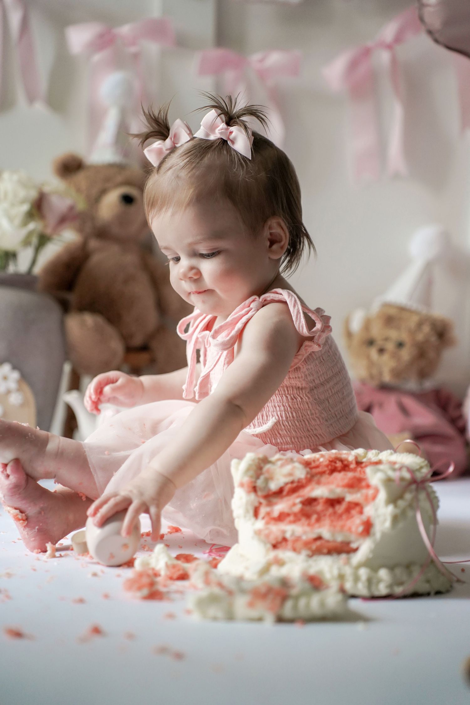 Baby in pink dress having fun smashing and playing with a pink cake during cake smash photo session with teddy bears nearby.