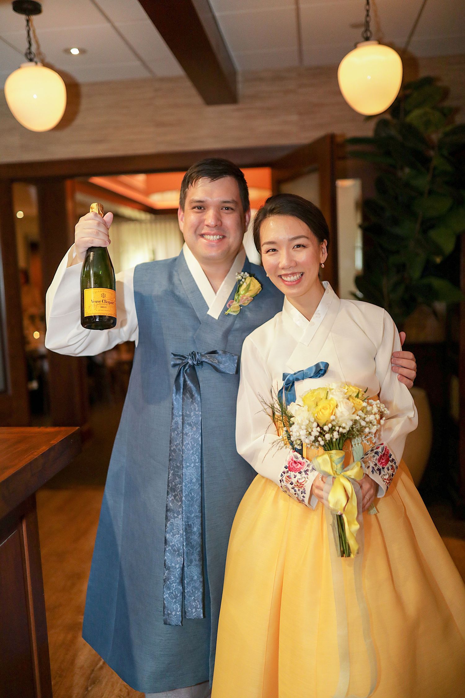A couple in traditional Korean hanbok attire celebrates with champagne and yellow flowers at an indoor venue.