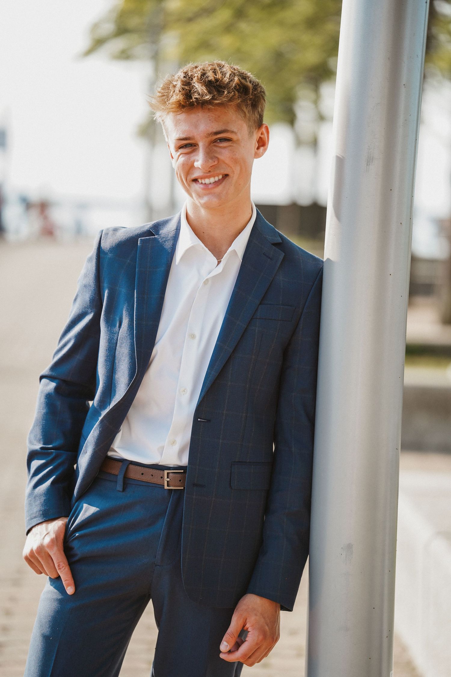Young man in navy blue business suit and white dress shirt smiling while leaning against pole outdoors.