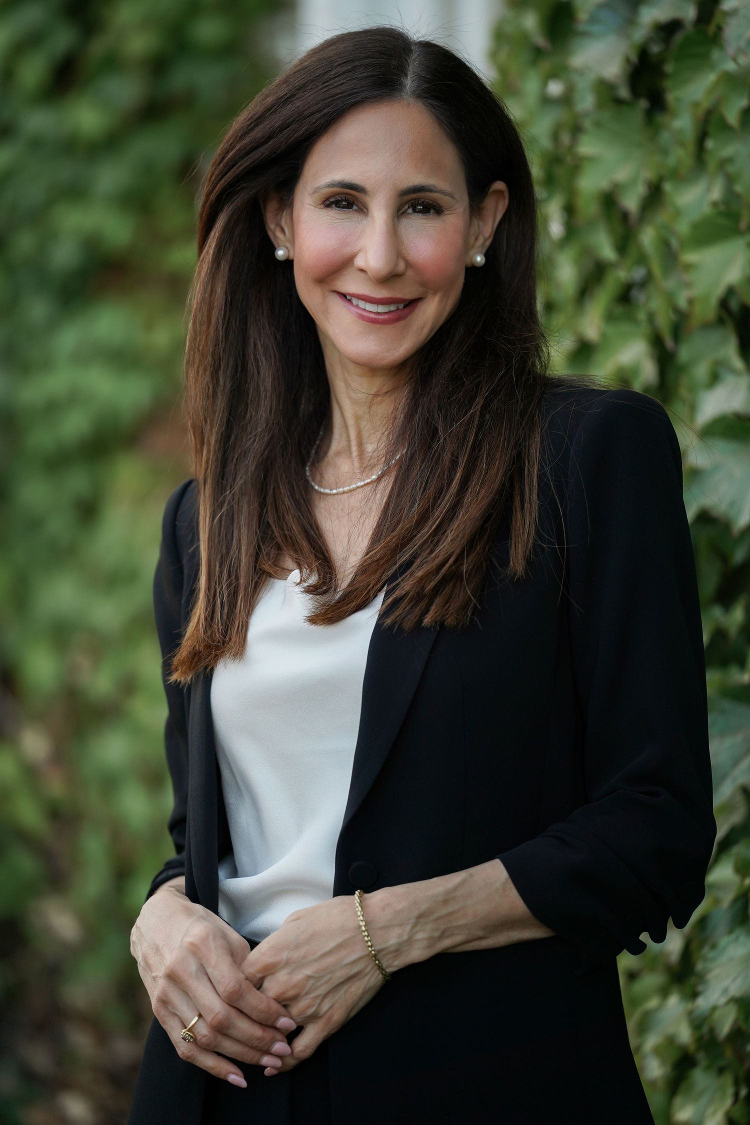 Professional headshot of someone in a black blazer and white top standing against a leafy green background.