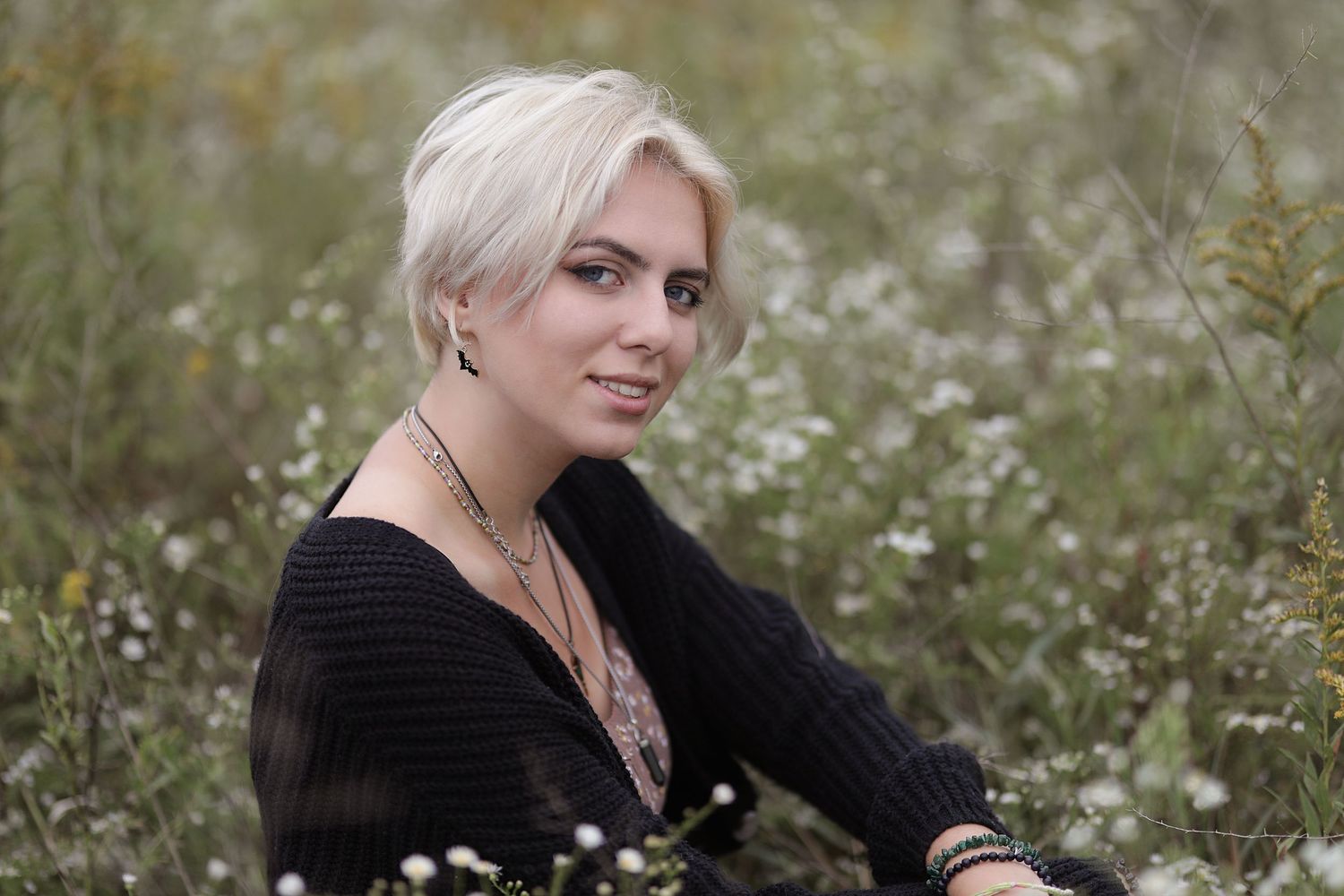 A model in a black sweater poses outdoors in a field of white wildflowers during a photoshoot.