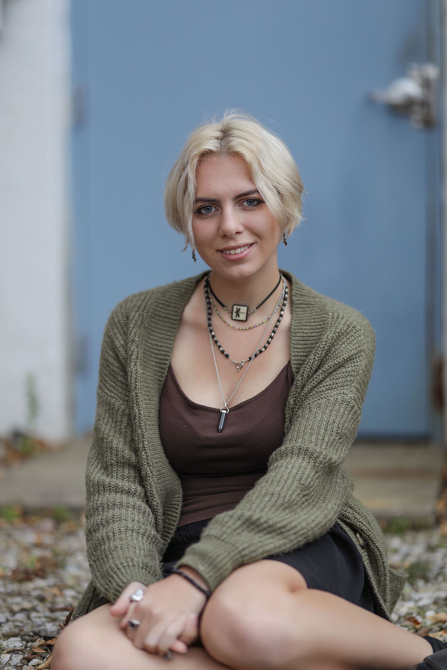 A person in a brown tank top and sage green cardigan poses outdoors wearing layered necklaces.