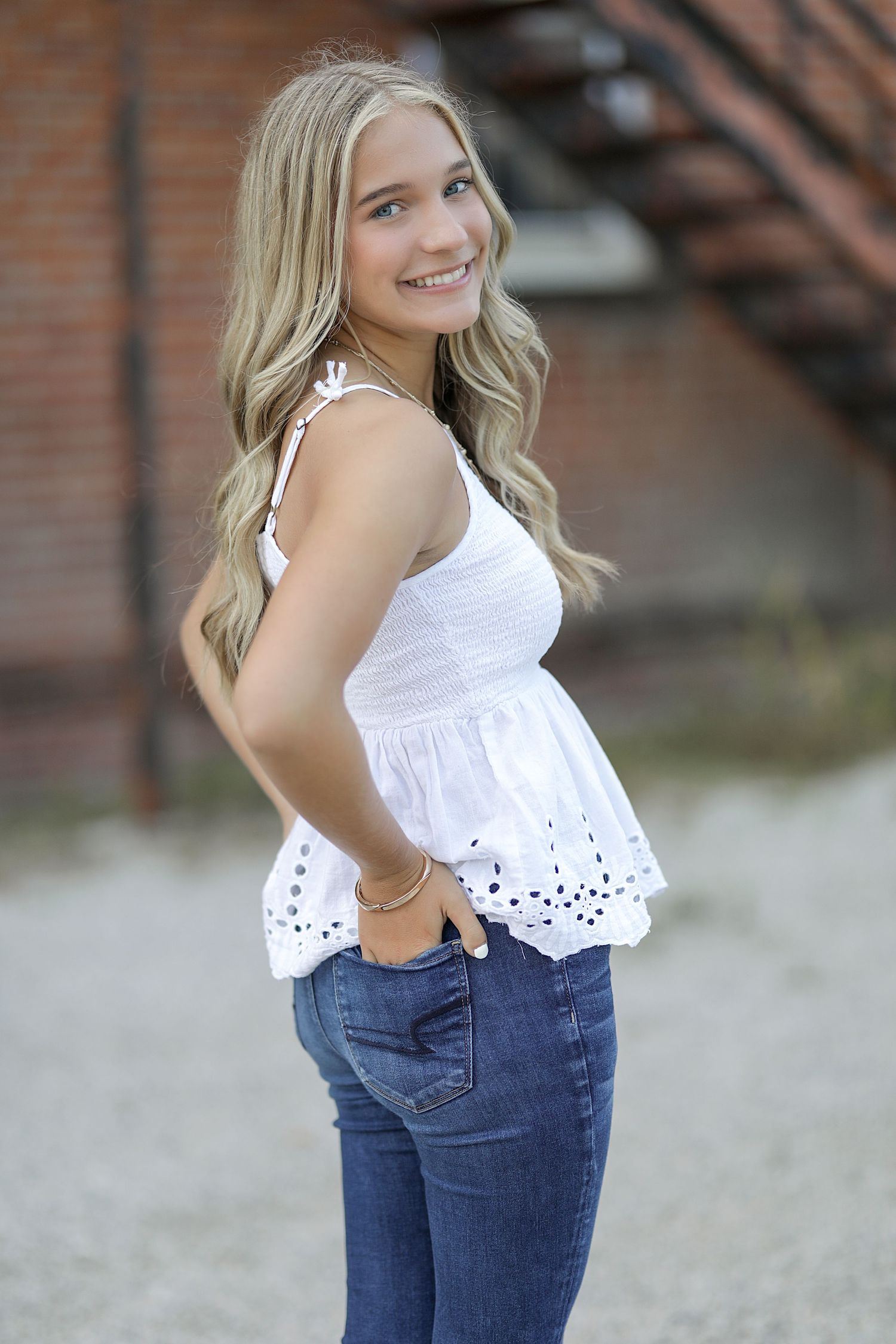 A bright white eyelet lace top pairs with blue jeans in an outdoor casual portrait against a brick wall background.
