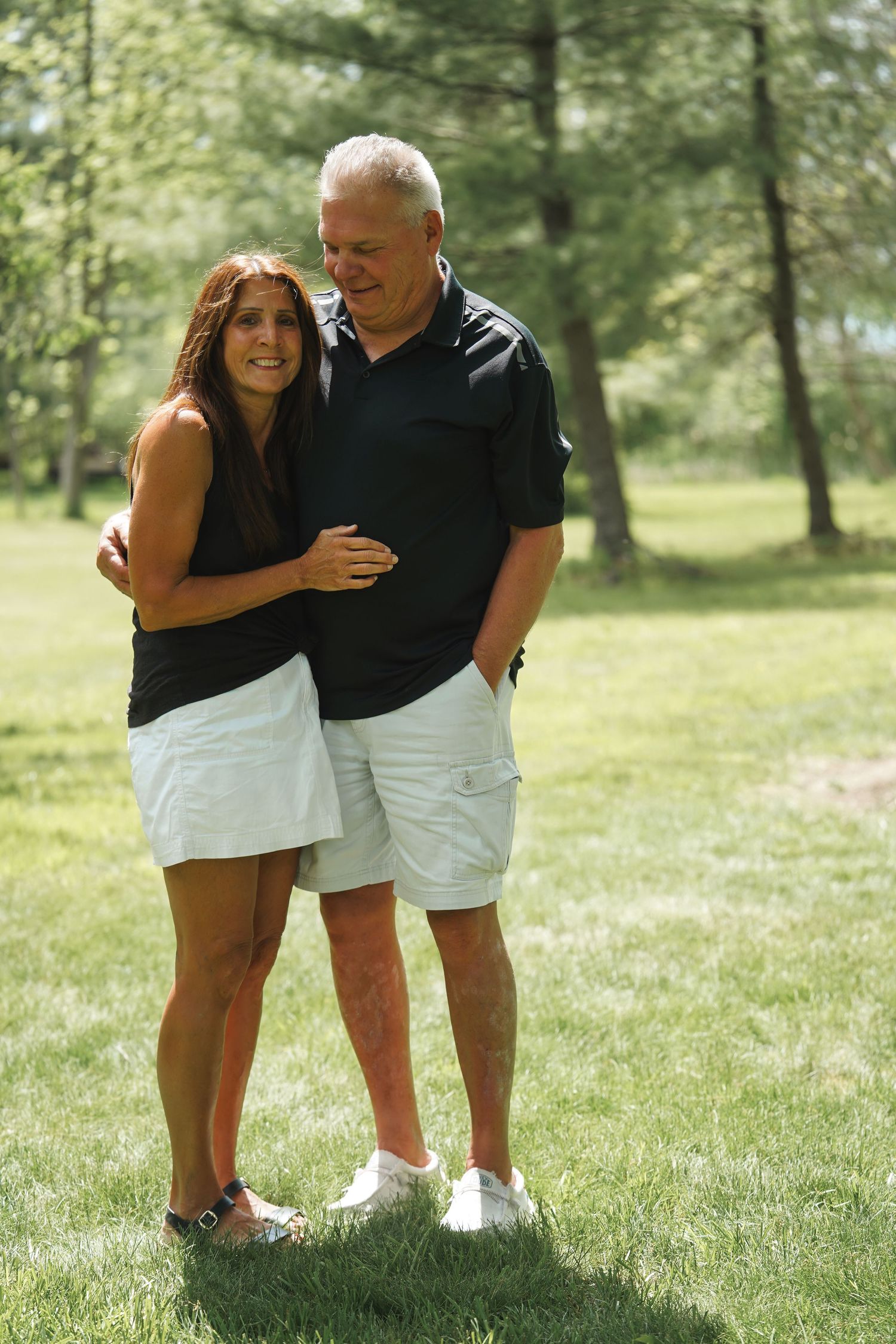 A couple wearing black shirts and white shorts embrace while standing on grass in a sunlit park setting.