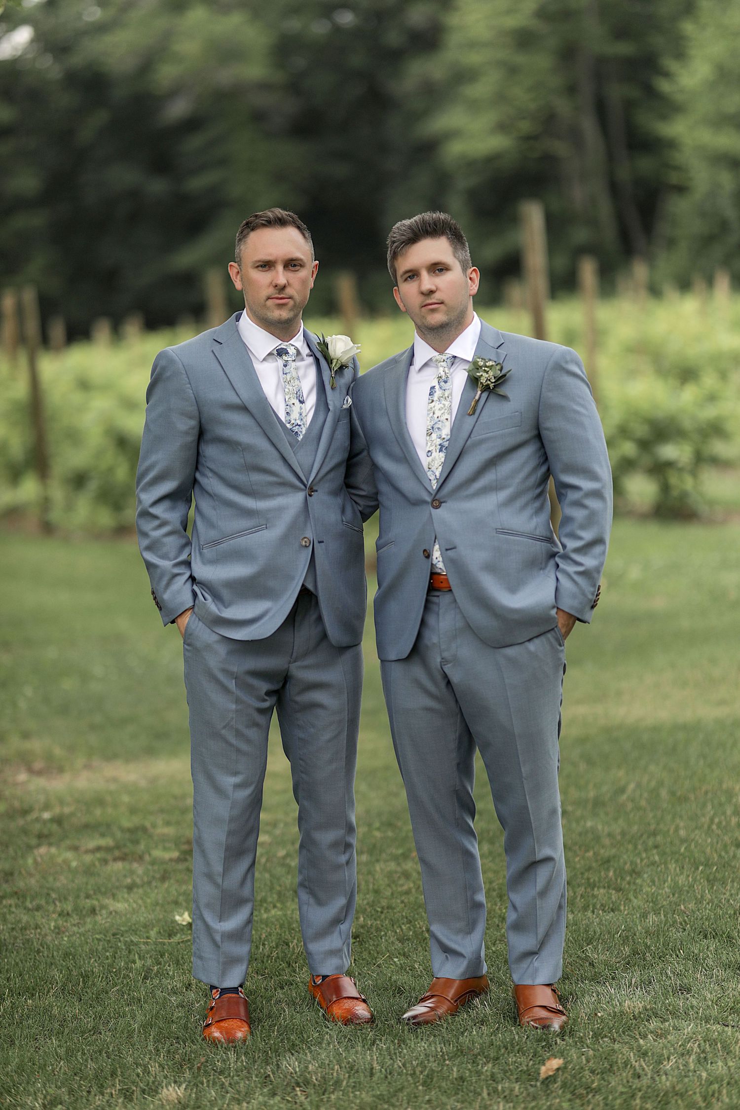 Two groomsmen in light blue suits and brown shoes pose casually on grass.