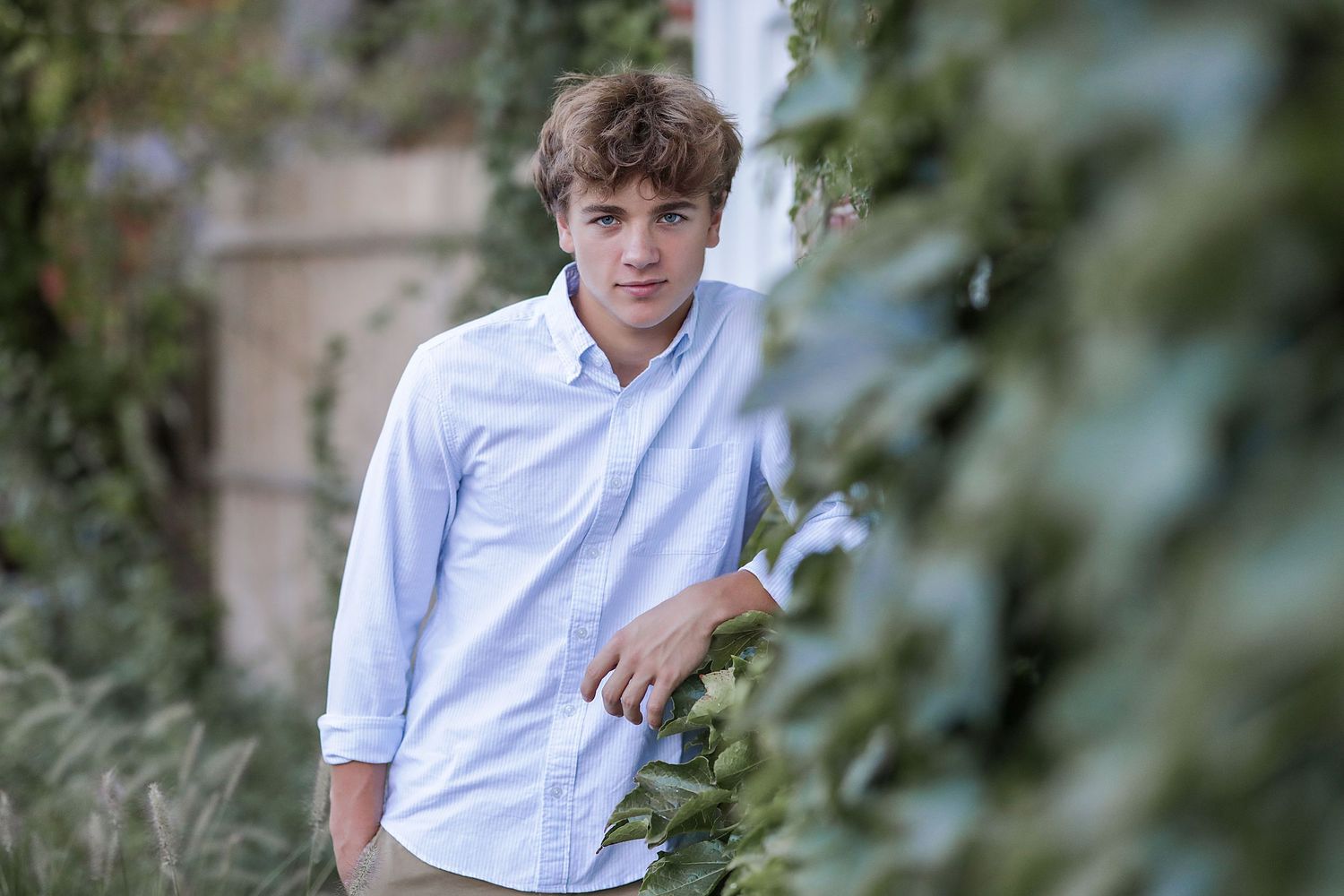 A person in a light blue button-down shirt stands outdoors among green foliage on a sunny day.
