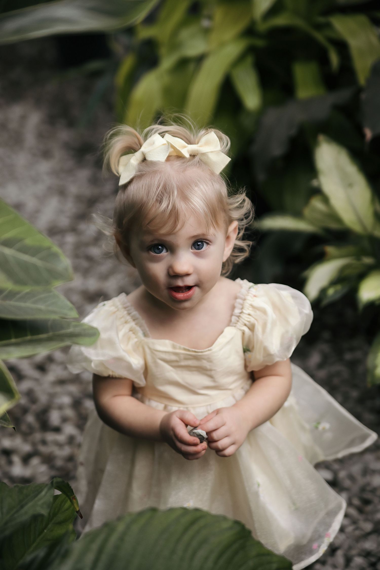 A small child in a cream-colored dress with puff sleeves poses among green foliage in a garden setting.