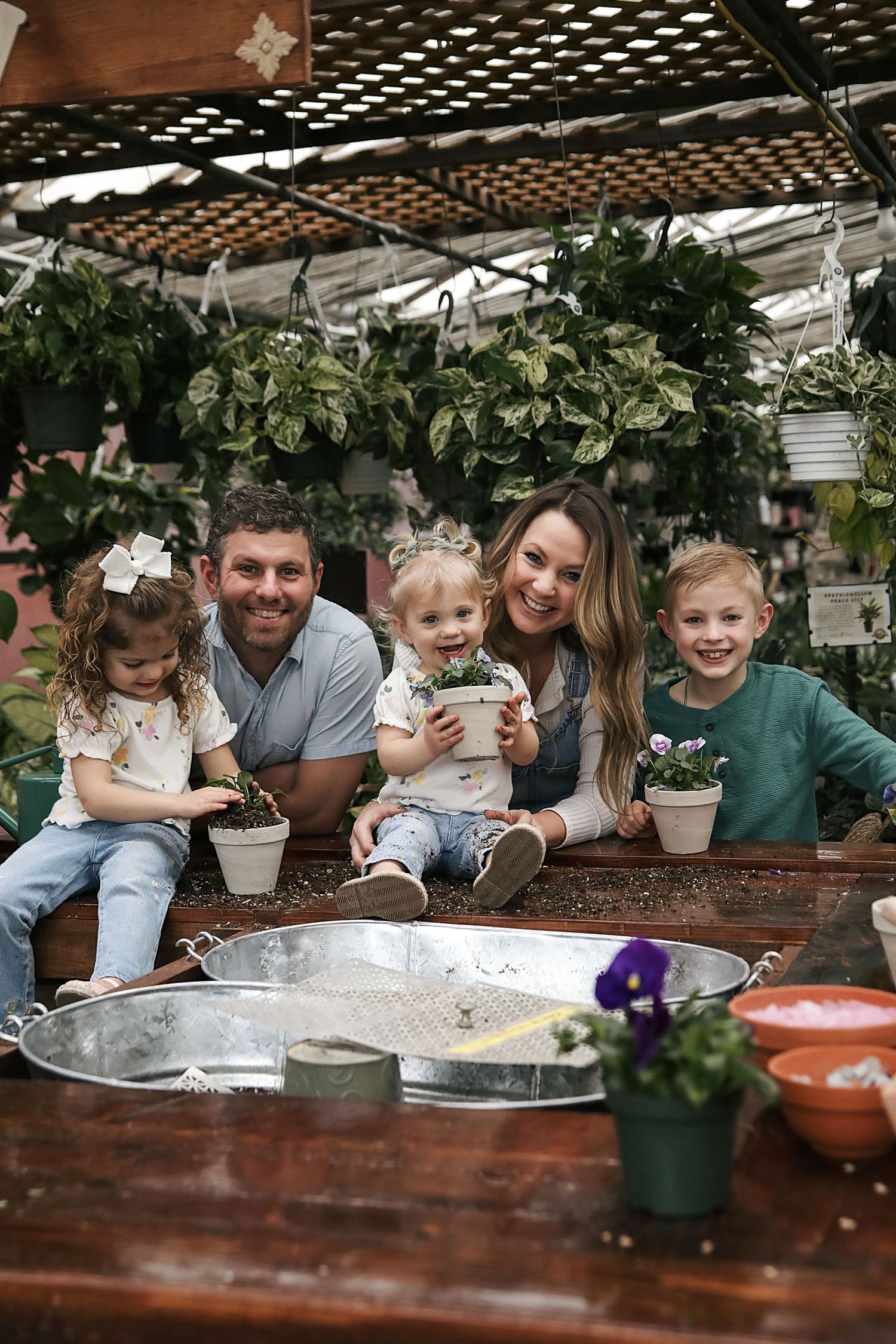A family sits together at a wooden table with potted plants in a greenhouse or garden setting.