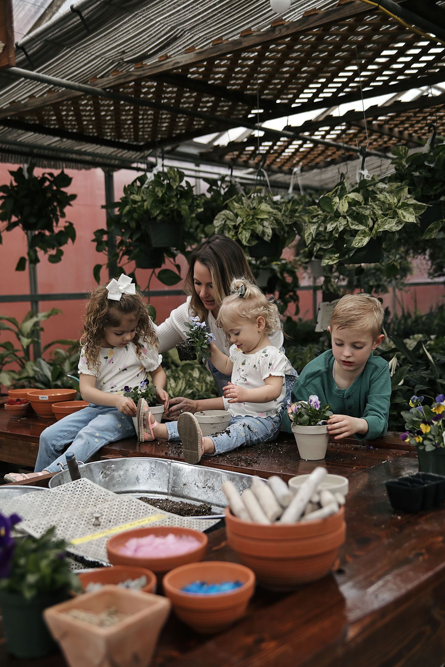 A small group gathered around a wooden table in a greenhouse with terra cotta pots and gardening supplies for planting.