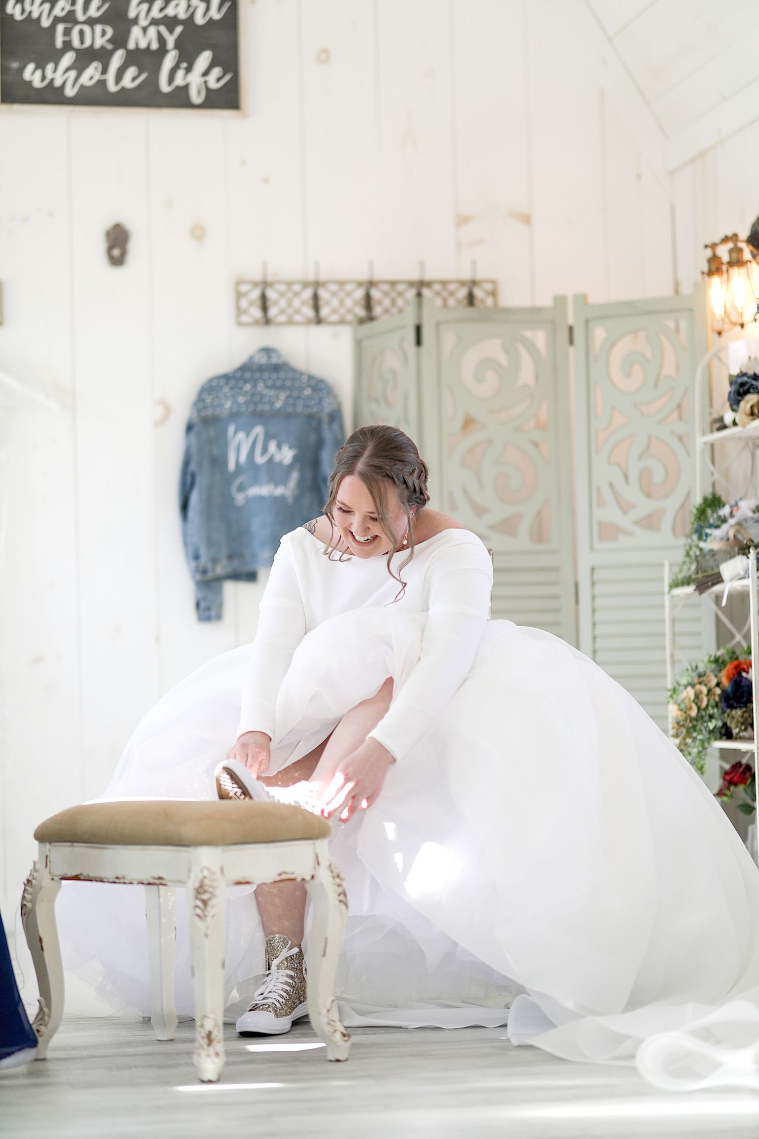 Bride in white dress and sneakers playfully bends down to adjust her shoe in a rustic wedding setting. Cleveland wedding photographer.