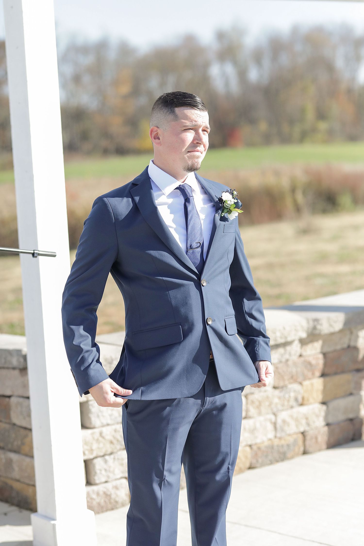 Groom in navy suit standing under white pergola during outdoor autumn ceremony.