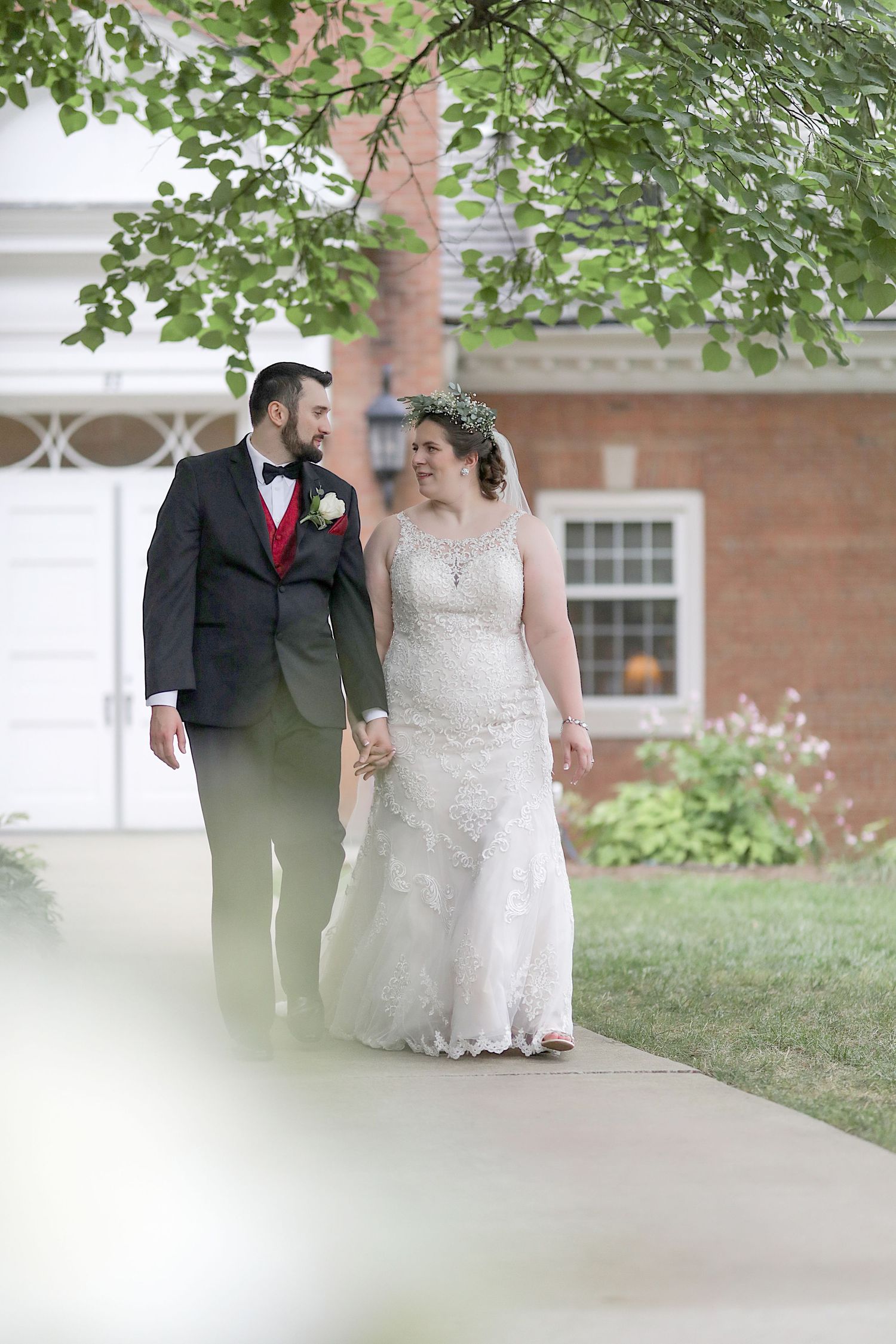 A bride and groom share a romantic moment together under green tree branches near a brick building.