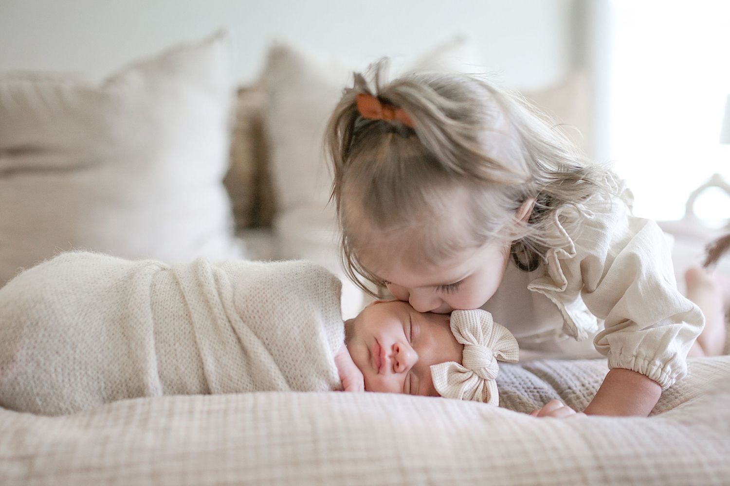 A young child in a white dress leans down to kiss a sleeping baby on a soft white linen bed.