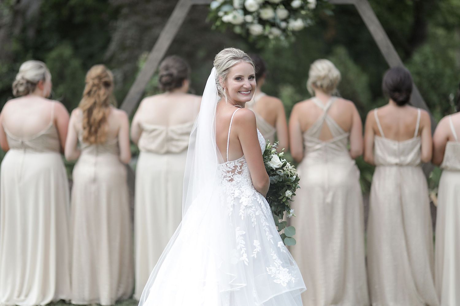 Row of bridesmaids in cream colored dresses standing in line at outdoor wedding ceremony.