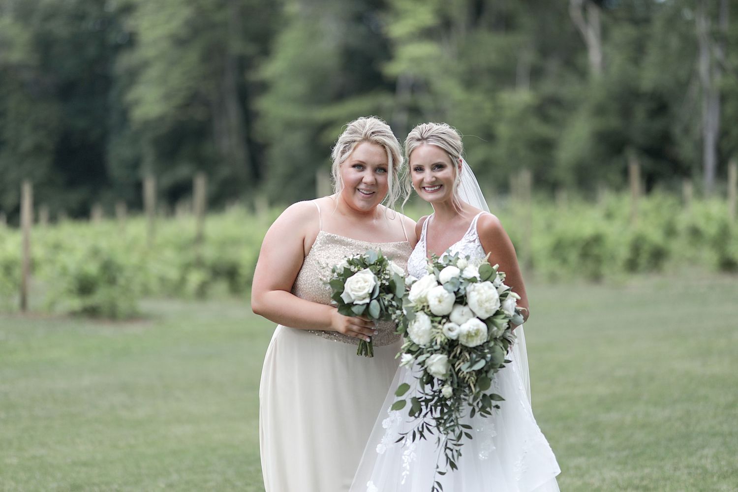 Bridesmaids in white dresses holding elegant bouquets pose together on grassy field.