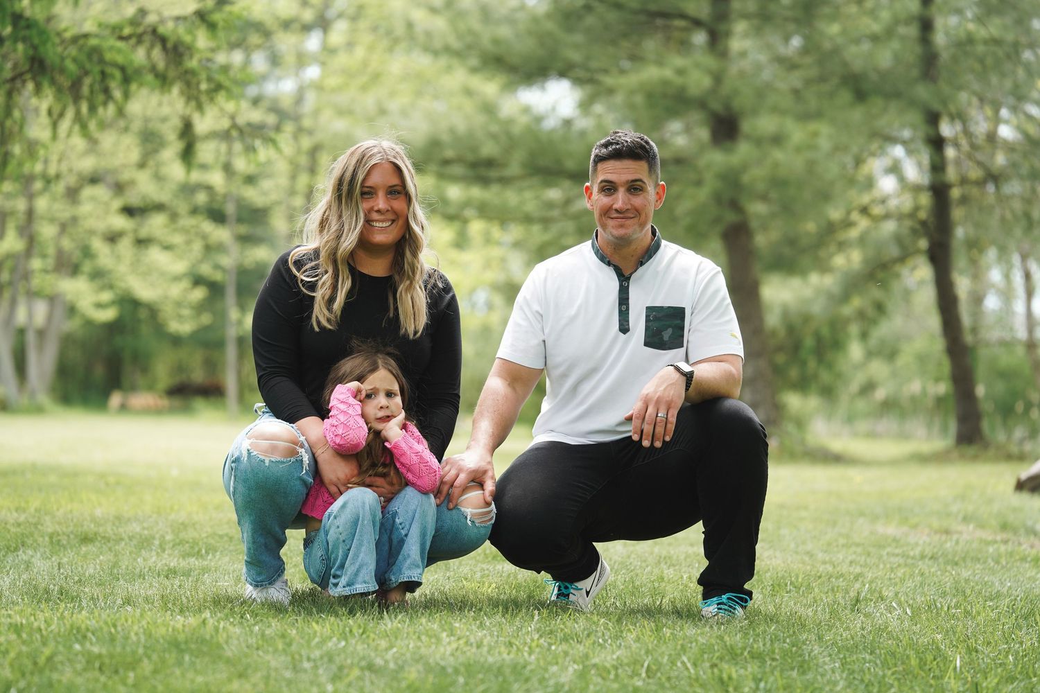 A family poses together on green grass in a wooded outdoor setting during a photo session.