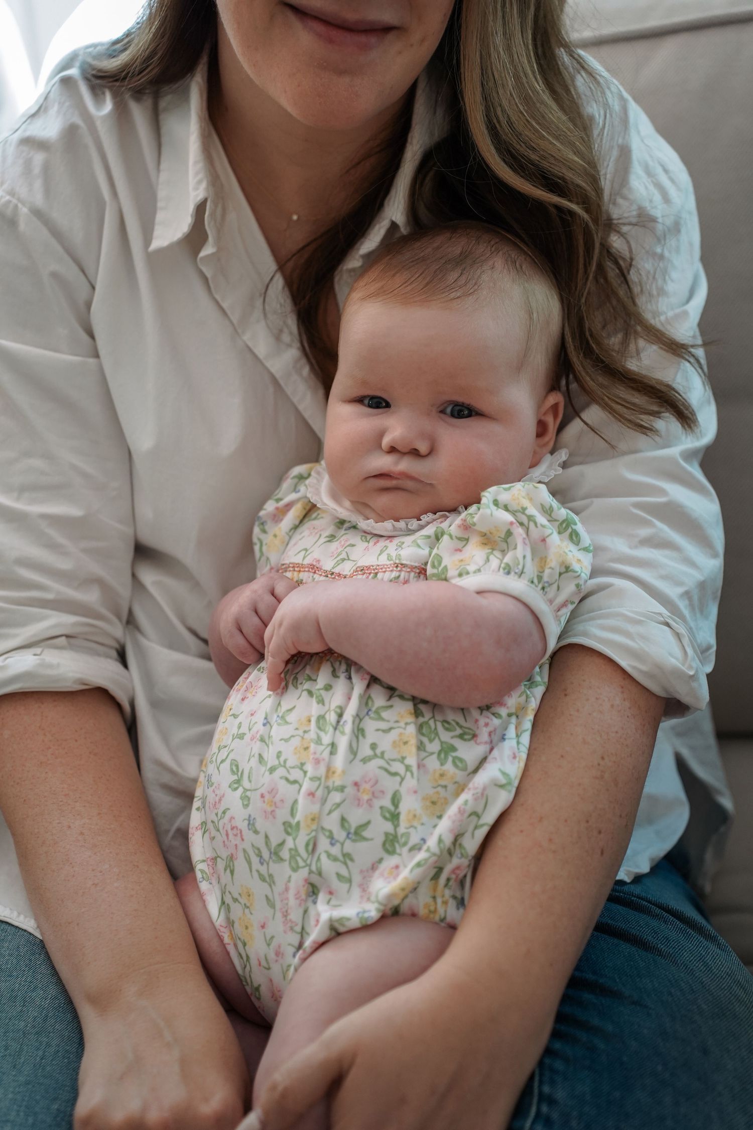 A baby wearing a floral romper sits on someone's lap wearing a white button-down shirt and jeans.