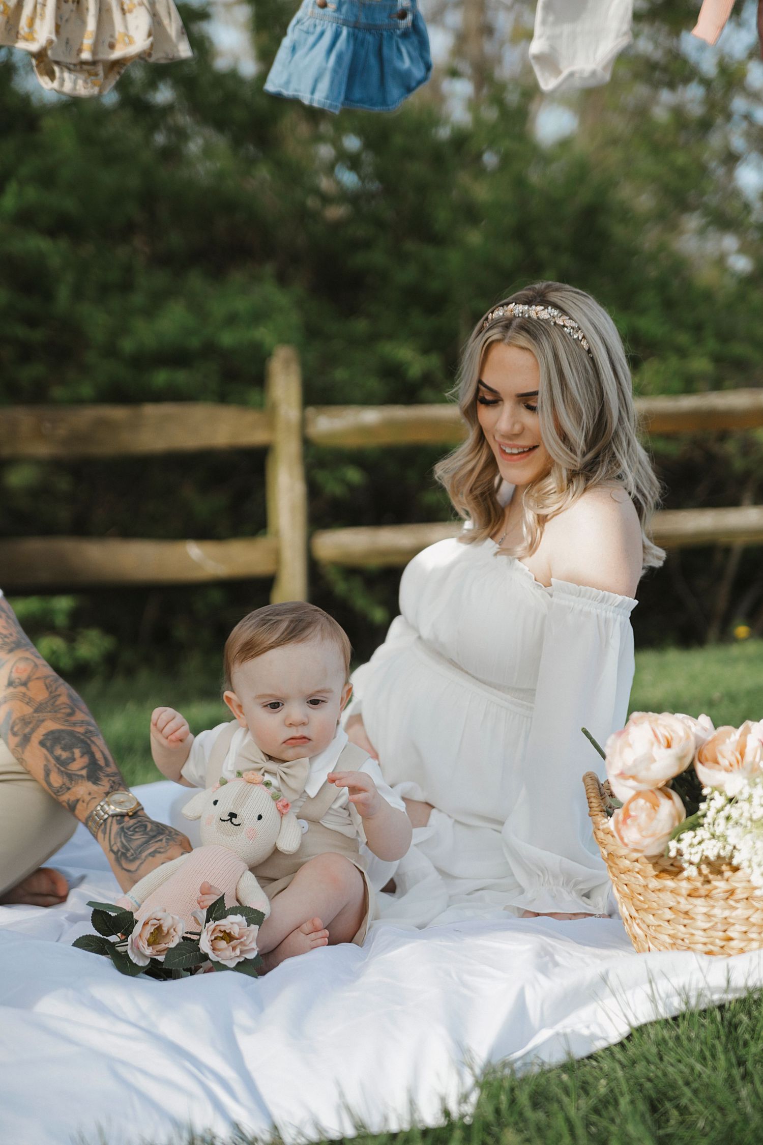 Two people enjoy a picnic on a white blanket outdoors with a basket of flowers and baby clothes hanging above.