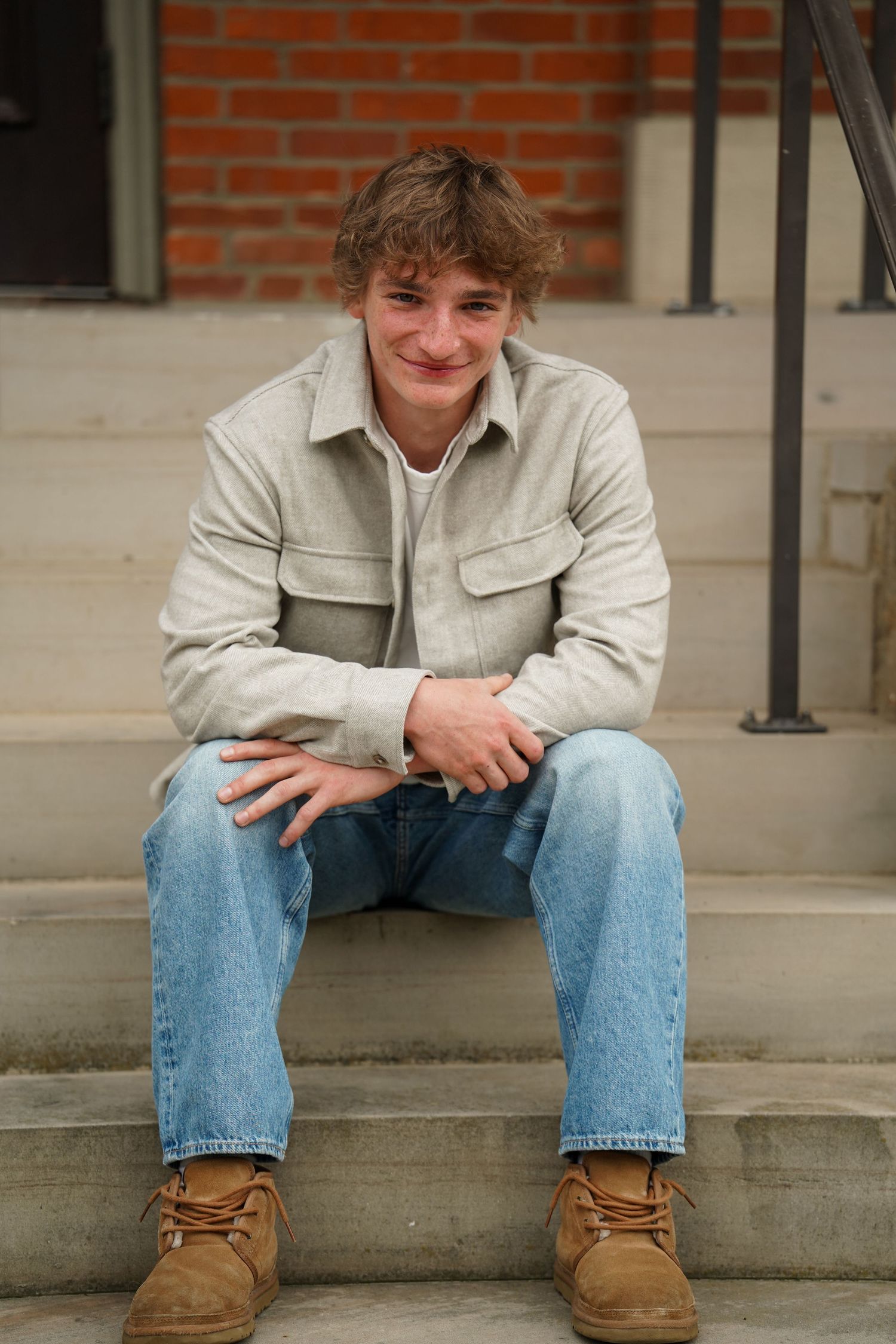 Someone in casual clothing sits on concrete steps against a brick wall, wearing a beige jacket and jeans.