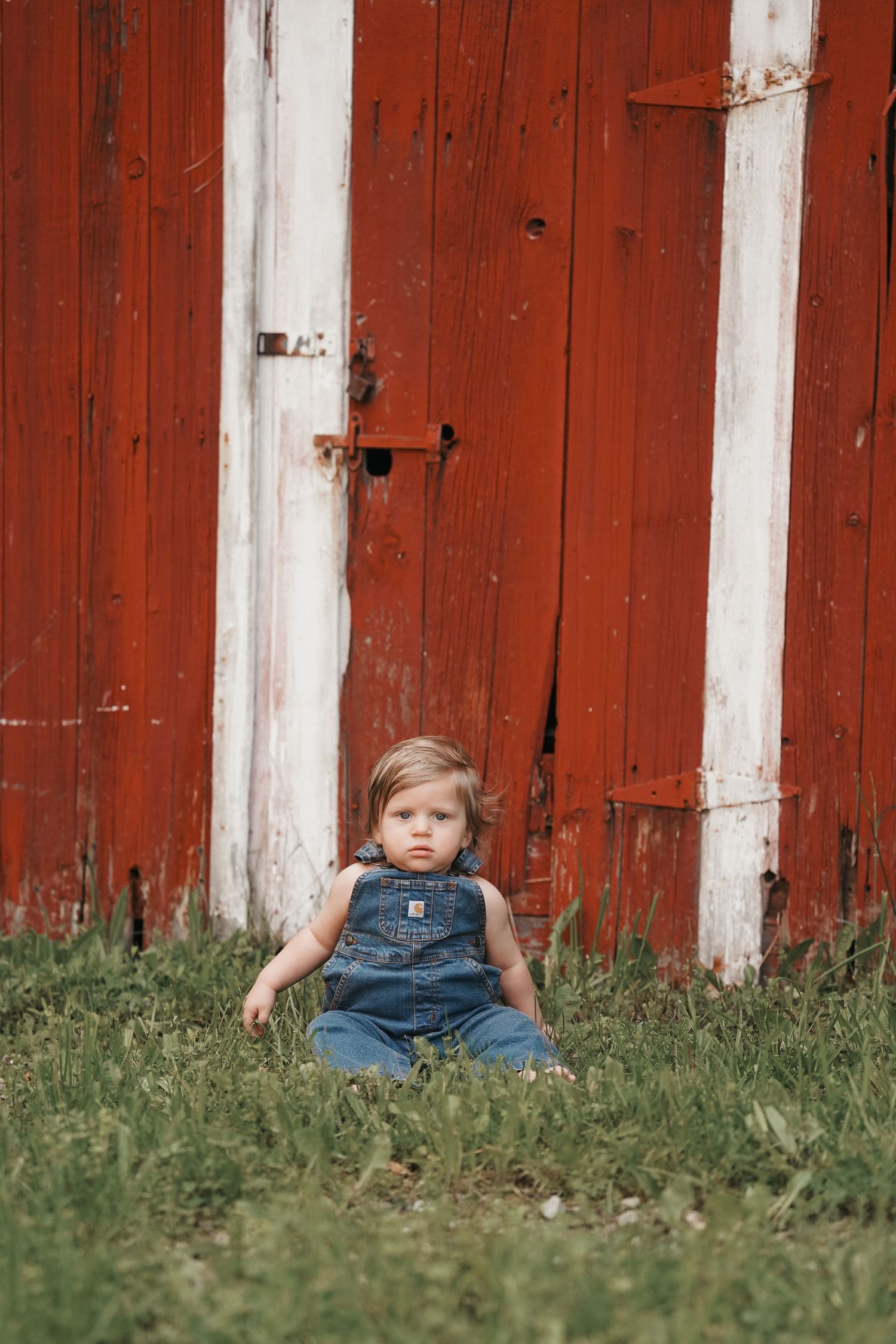 Baby in denim overalls sits in green grass against a rustic red barn wall in a warm summer scene.