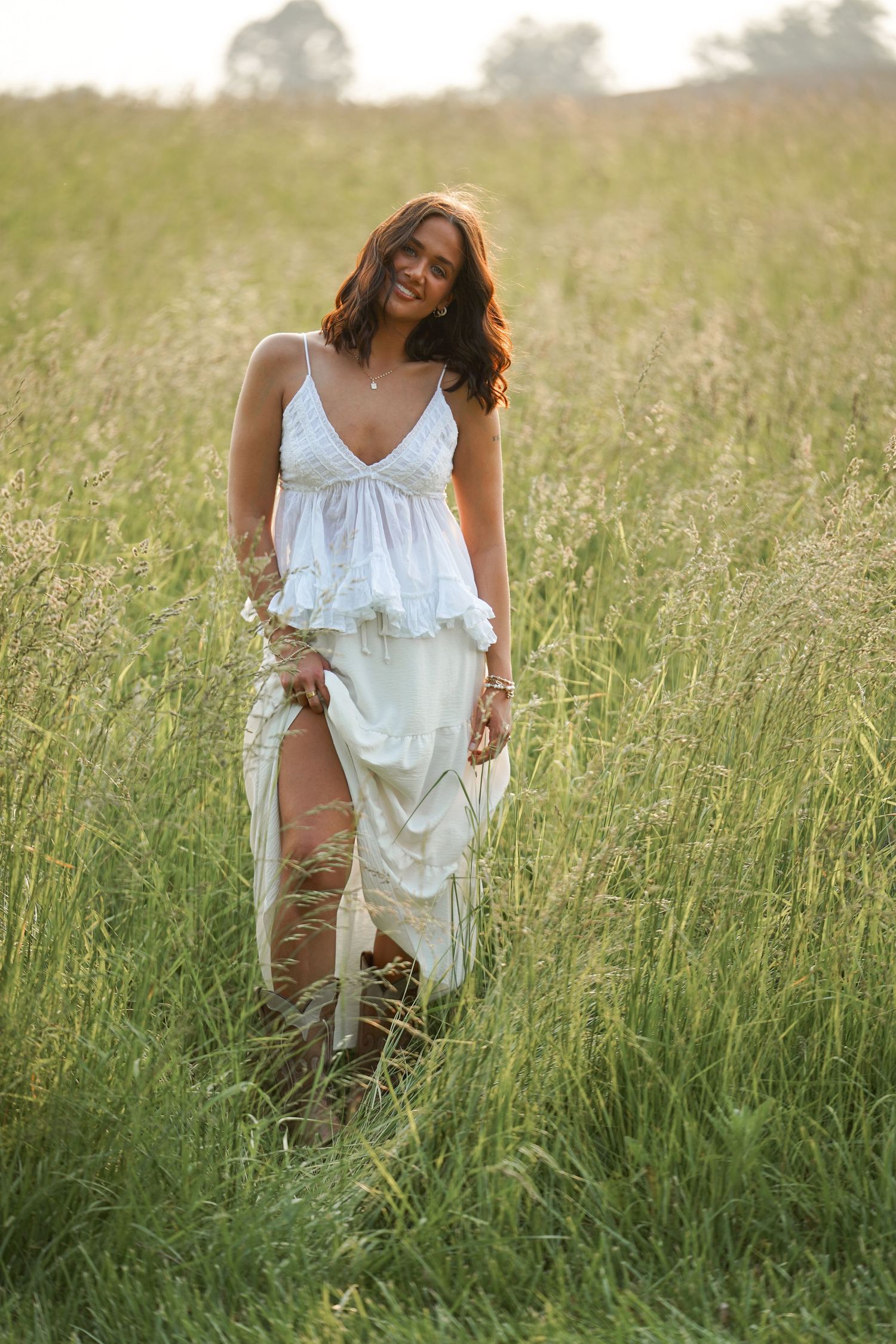 A figure in a flowing white dress walks through tall grass in a serene outdoor setting at golden hour.