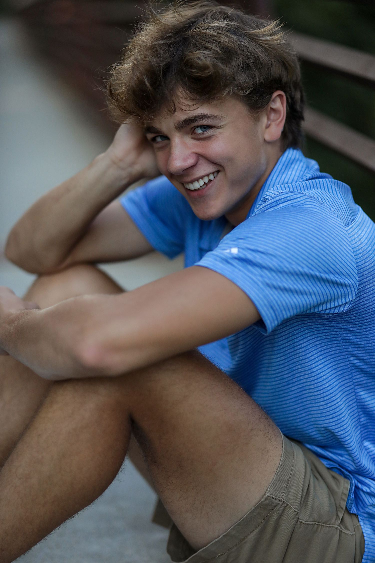 Smiling person wearing a blue striped polo shirt and khaki shorts sitting outdoors on a summer day.