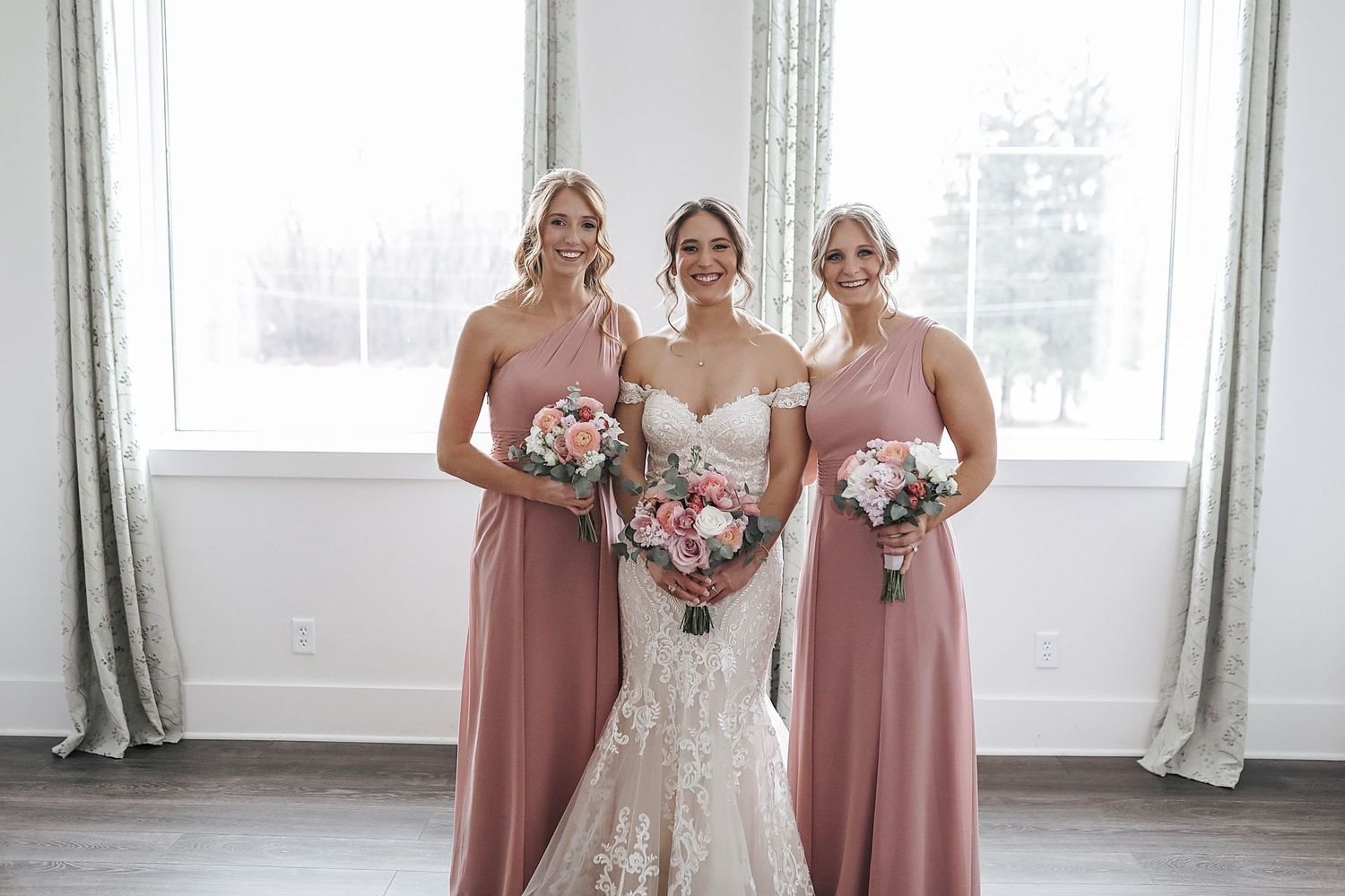 A bride in a white gown stands with bridesmaids wearing dusty rose dresses in a bright sunlit room.