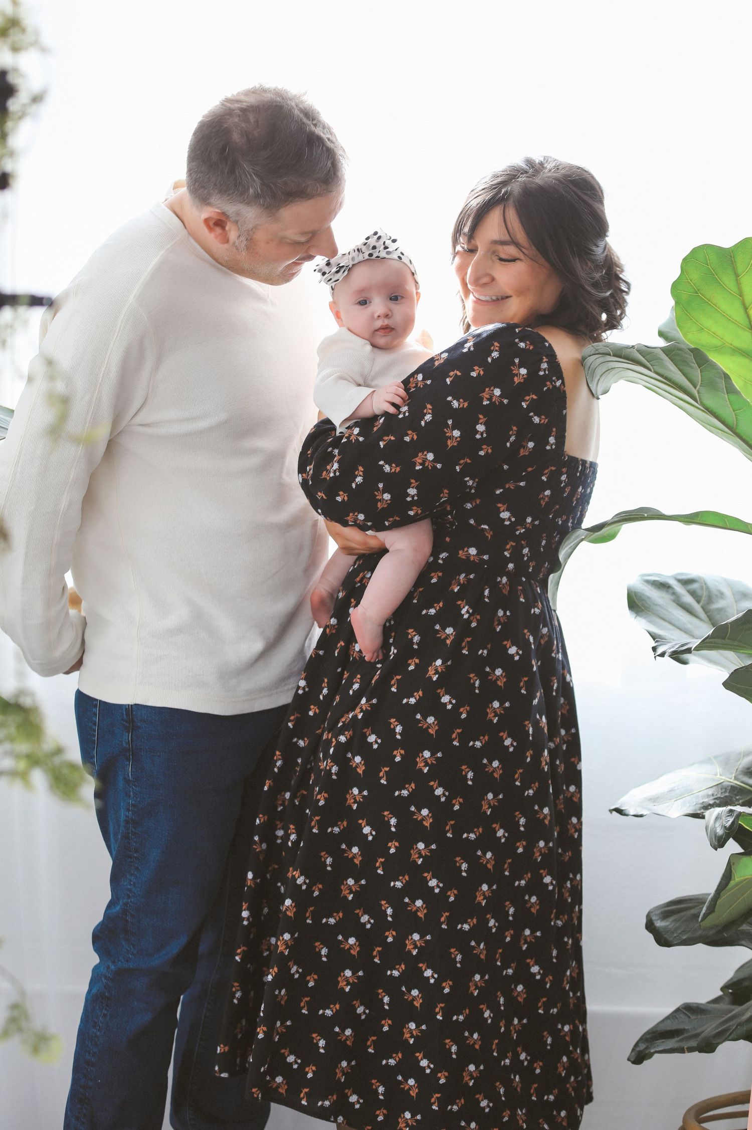 A couple in a white shirt and floral dress share tender moments with a baby near an indoor plant.