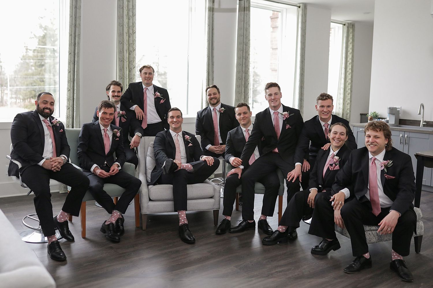A group of formally dressed groomsmen in black suits and pink ties gather in a modern bright room for wedding photos.