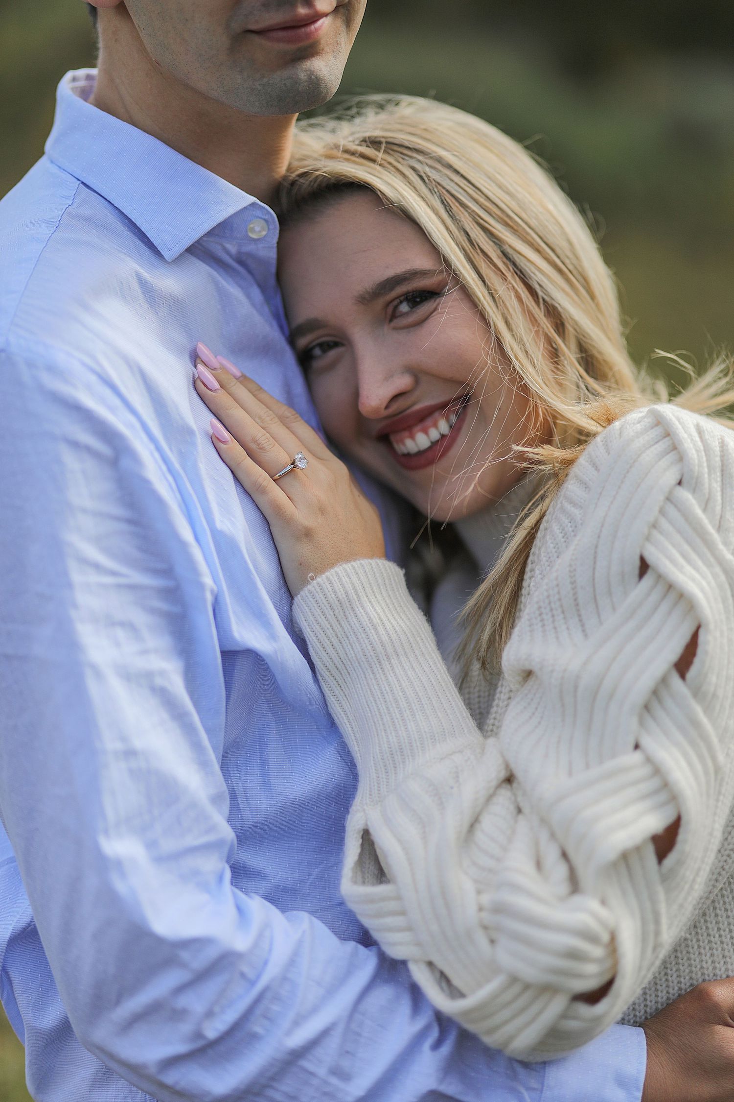 A couple embraces outdoors, with person wearing a cozy white sweater showing off an engagement ring against a blue shirt.