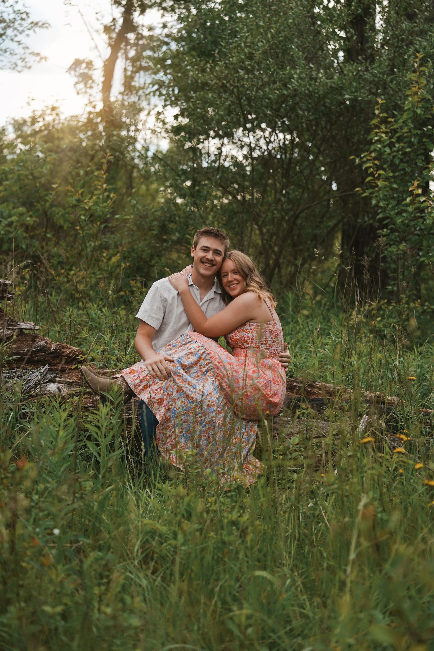A couple sits together on a fallen log in a lush green forest setting during golden hour.