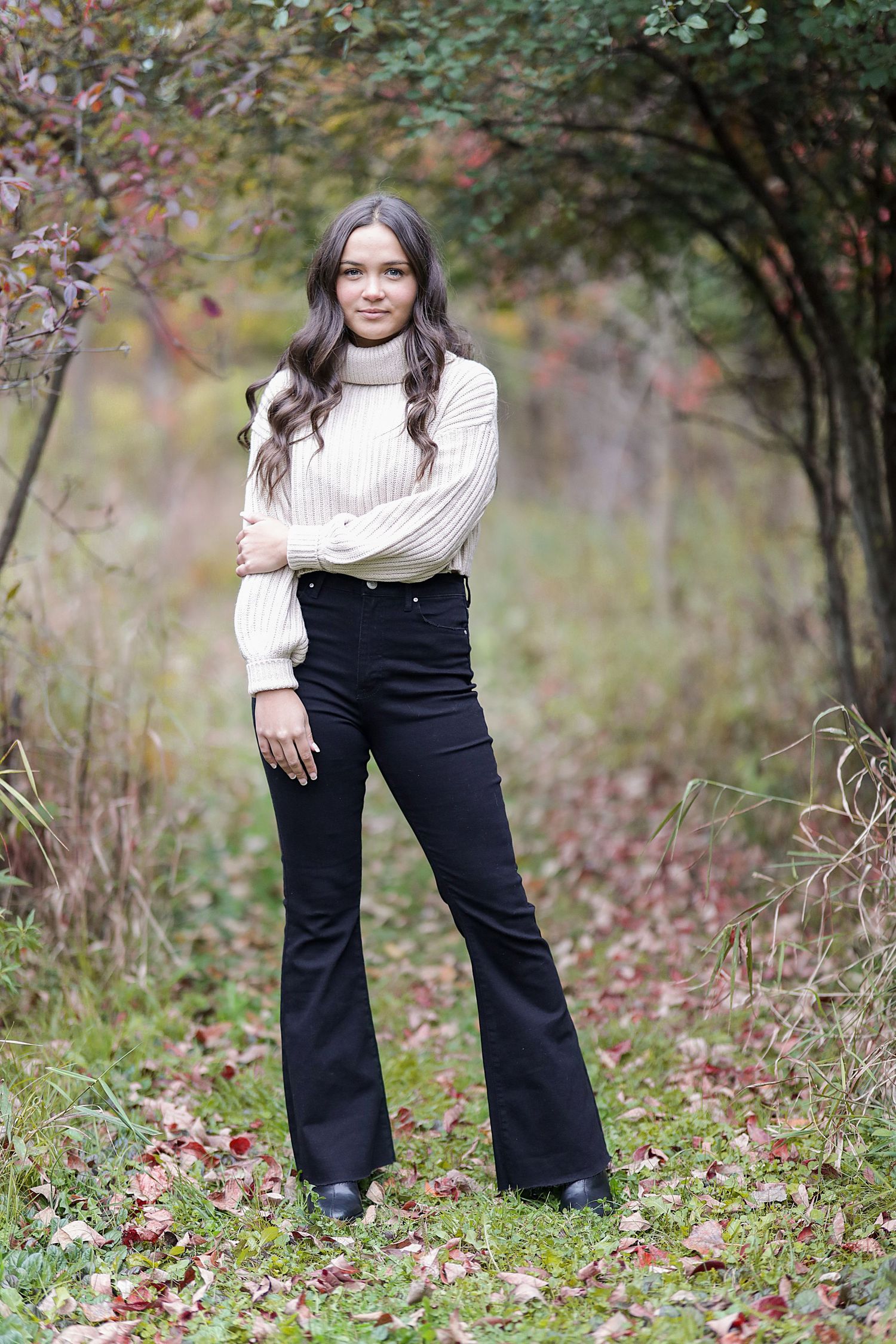 Person in white sweater and black overalls poses outdoors among autumn foliage during portrait shoot.