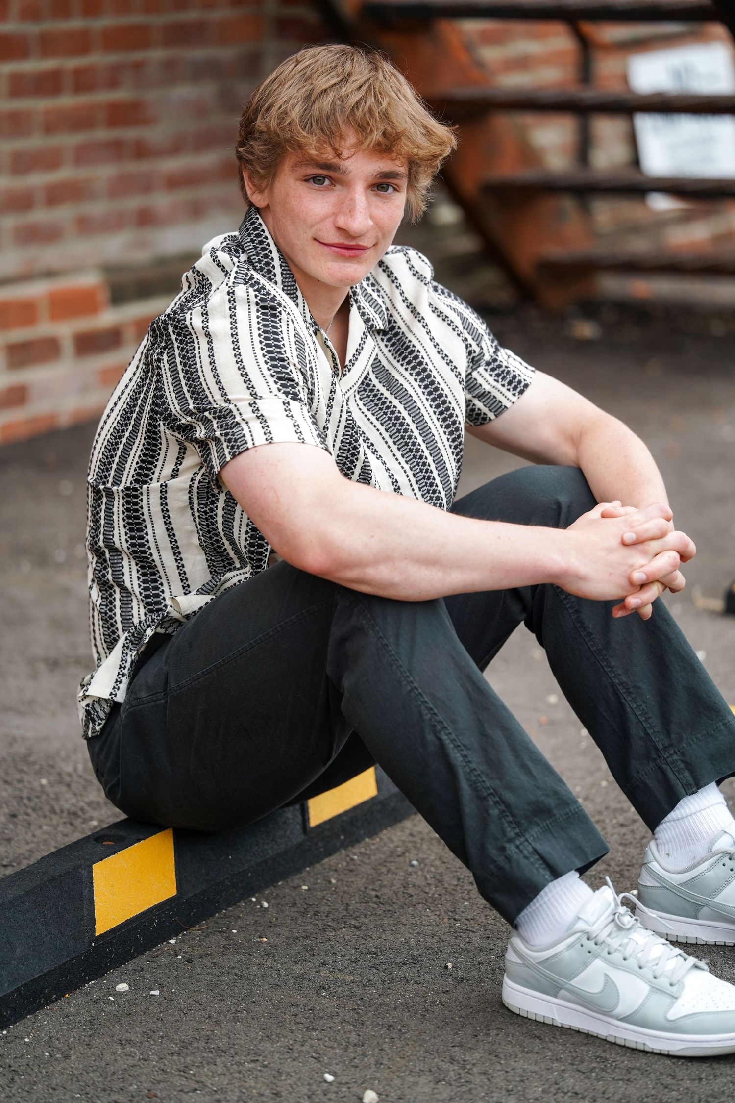Person in striped shirt and black pants sits casually against brick wall wearing grey sneakers.