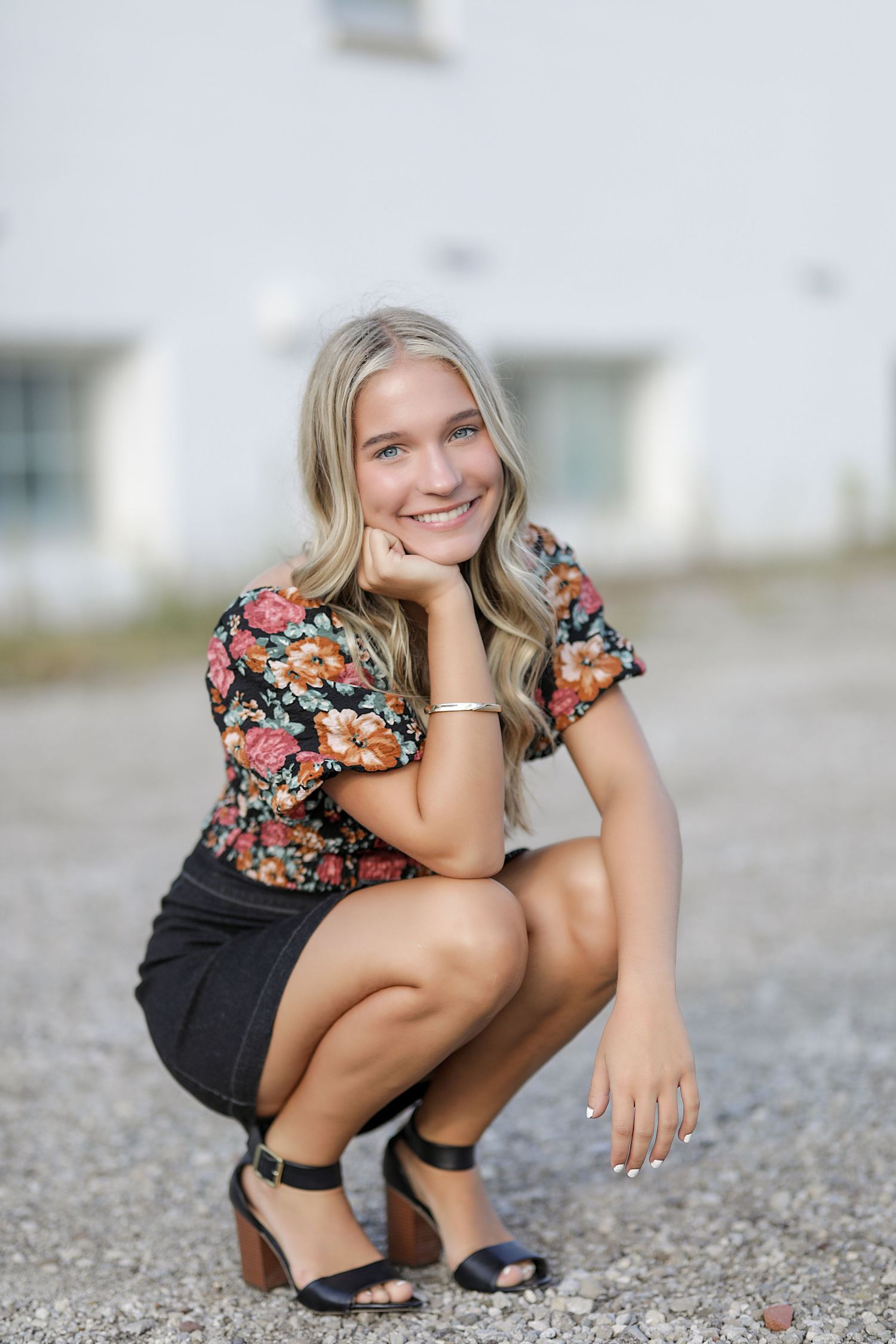 A person wearing a floral blouse and black shorts crouches on a gravel path in summer.