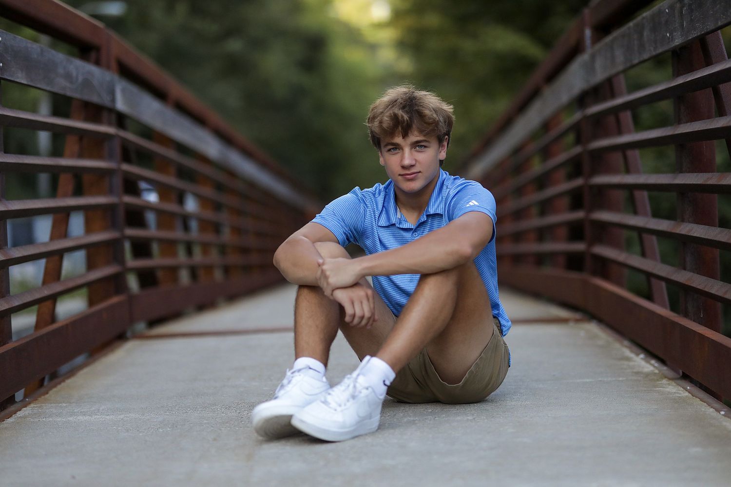 Someone in a blue shirt and khaki shorts sits casually on a wooden bridge on a summer afternoon.