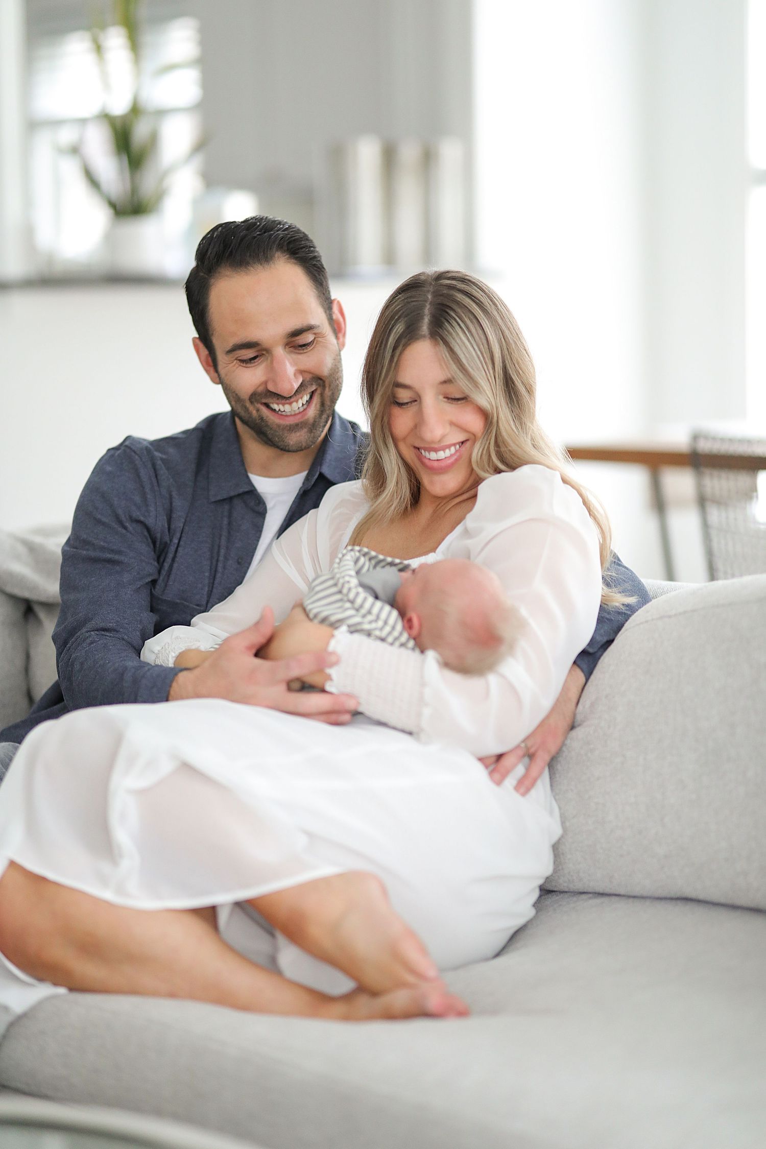 Happy parents smiling while holding their newborn baby on a white couch. Newborn photographer Chagrin Falls, Ohio.