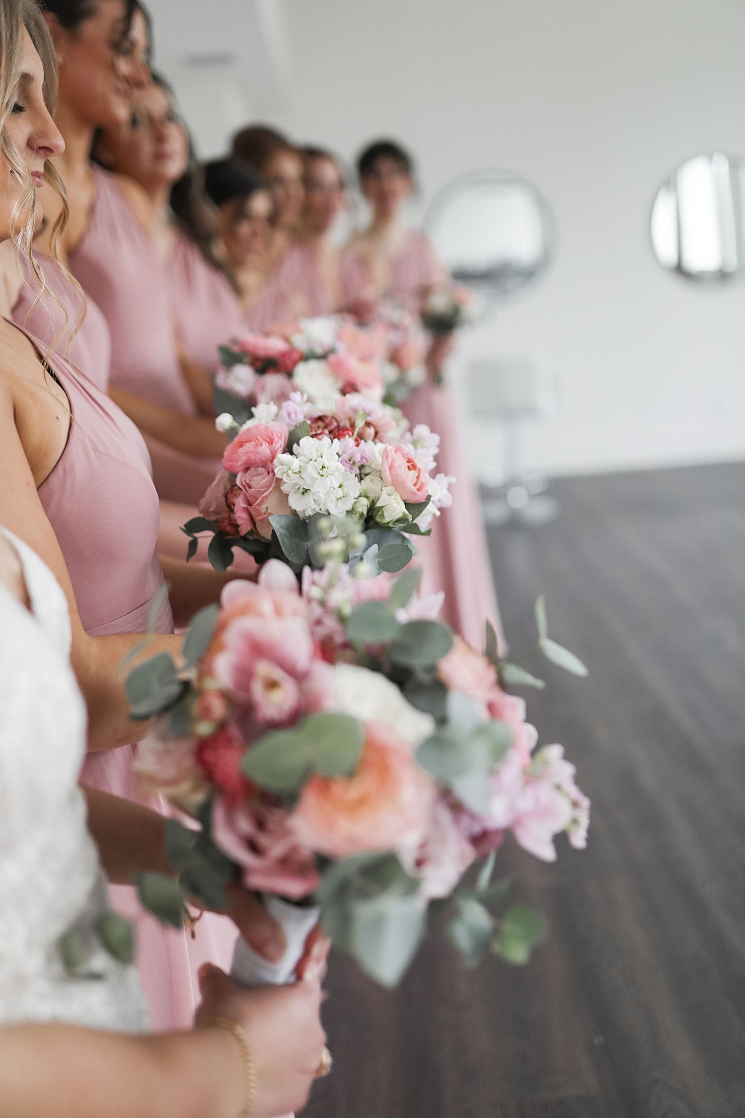 Bridesmaids in pink dresses hold elegant bouquets of pink and white roses with eucalyptus at a wedding ceremony.