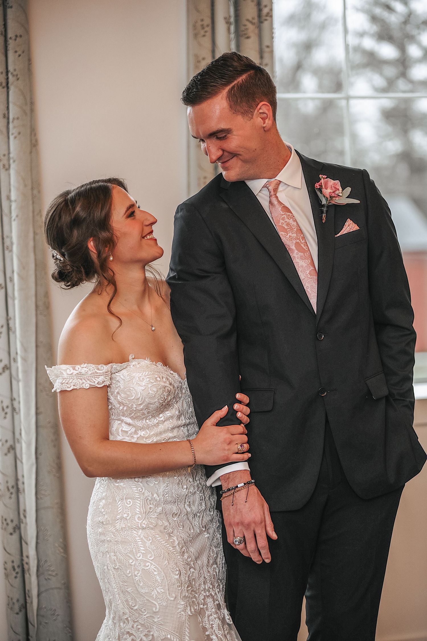 A wedding couple shares a candid laughing moment together in formal attire by large windows.