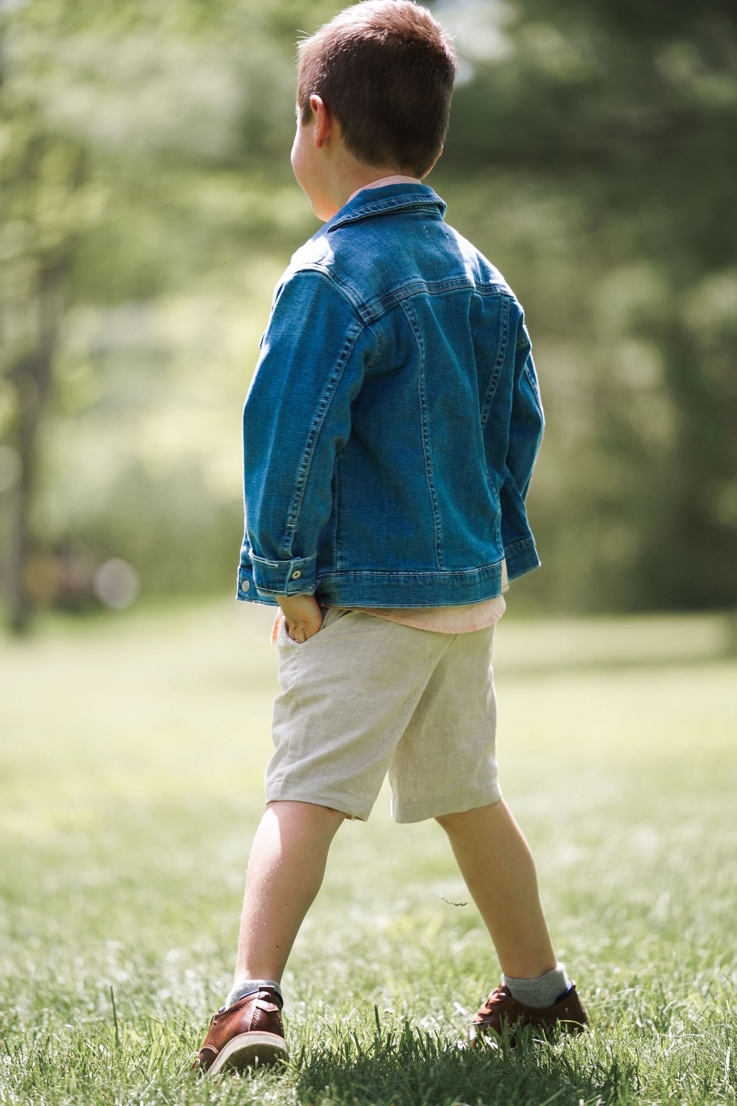 A child in a denim jacket and khaki shorts walking on green grass in a park setting.