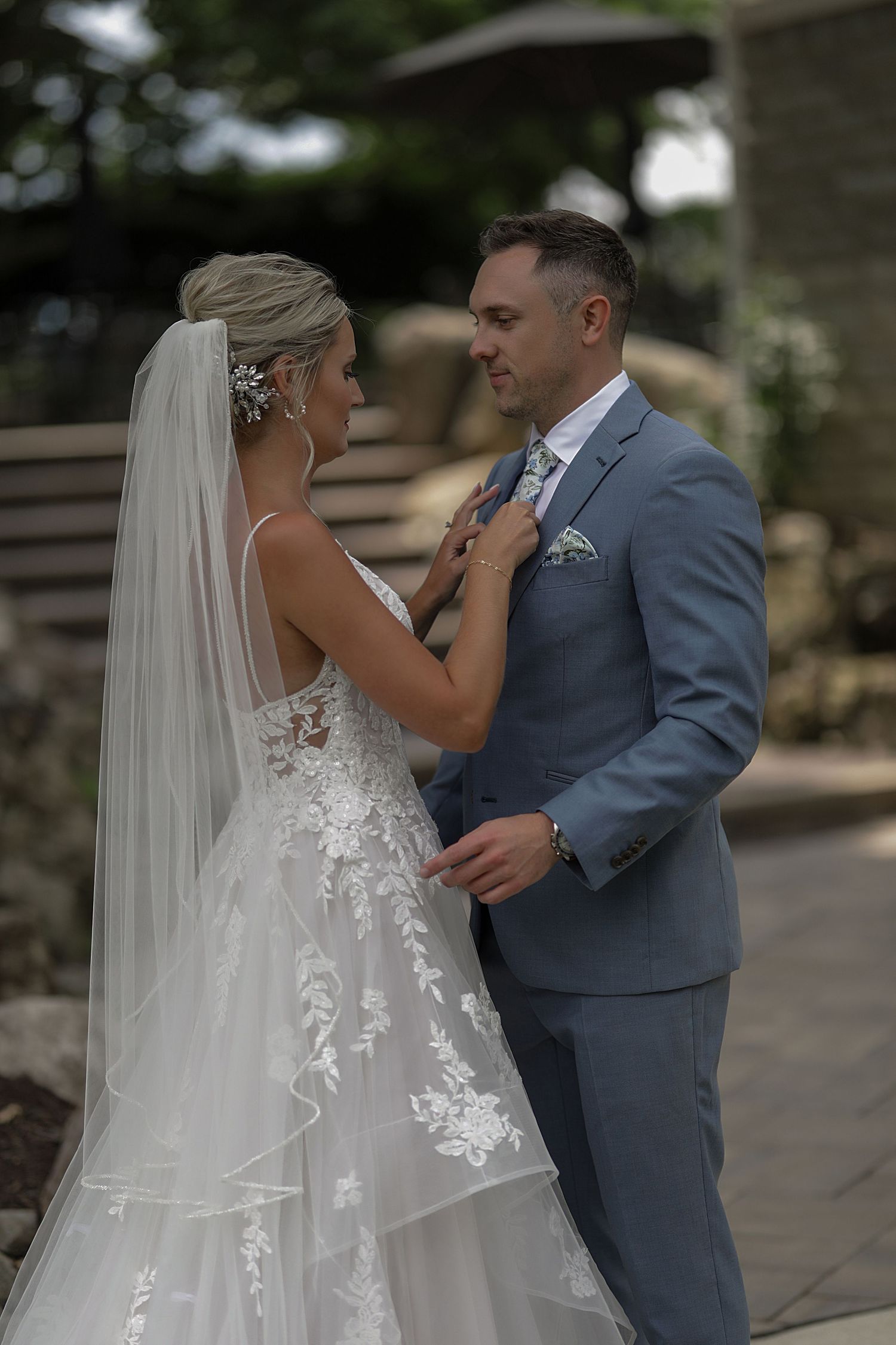 A tender moment between newlyweds as the bride in a lace gown adjusts the groom's grey suit tie outdoors.