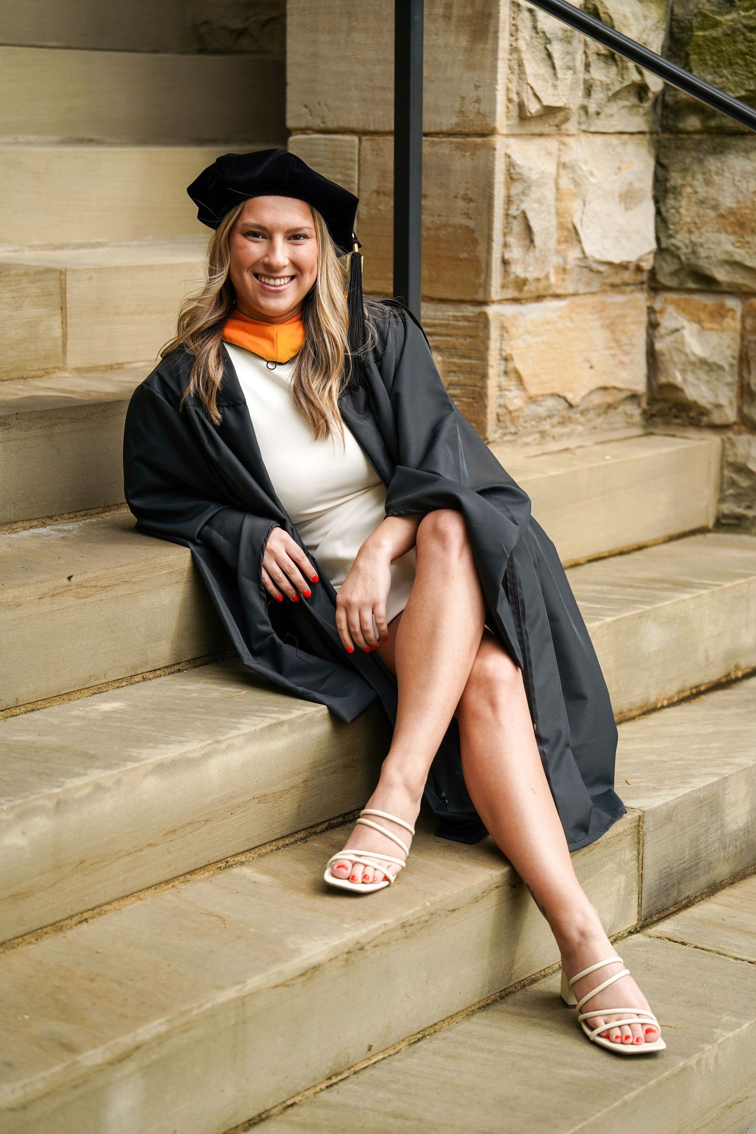Person in a black graduation gown sits on concrete steps wearing white sandals in a casual pose.