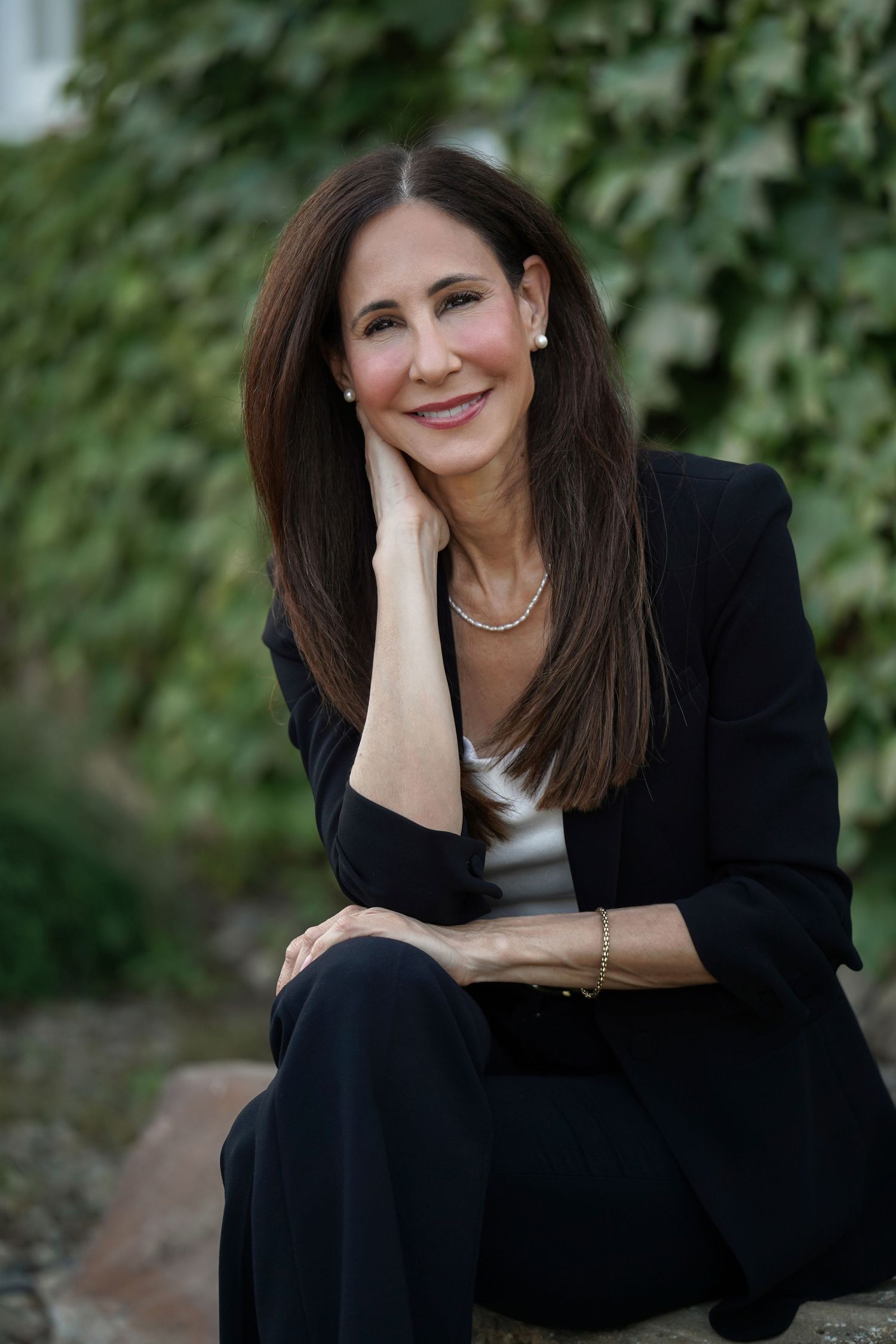 Professional business portrait outdoors with person wearing black blazer and white top sitting against green foliage background.