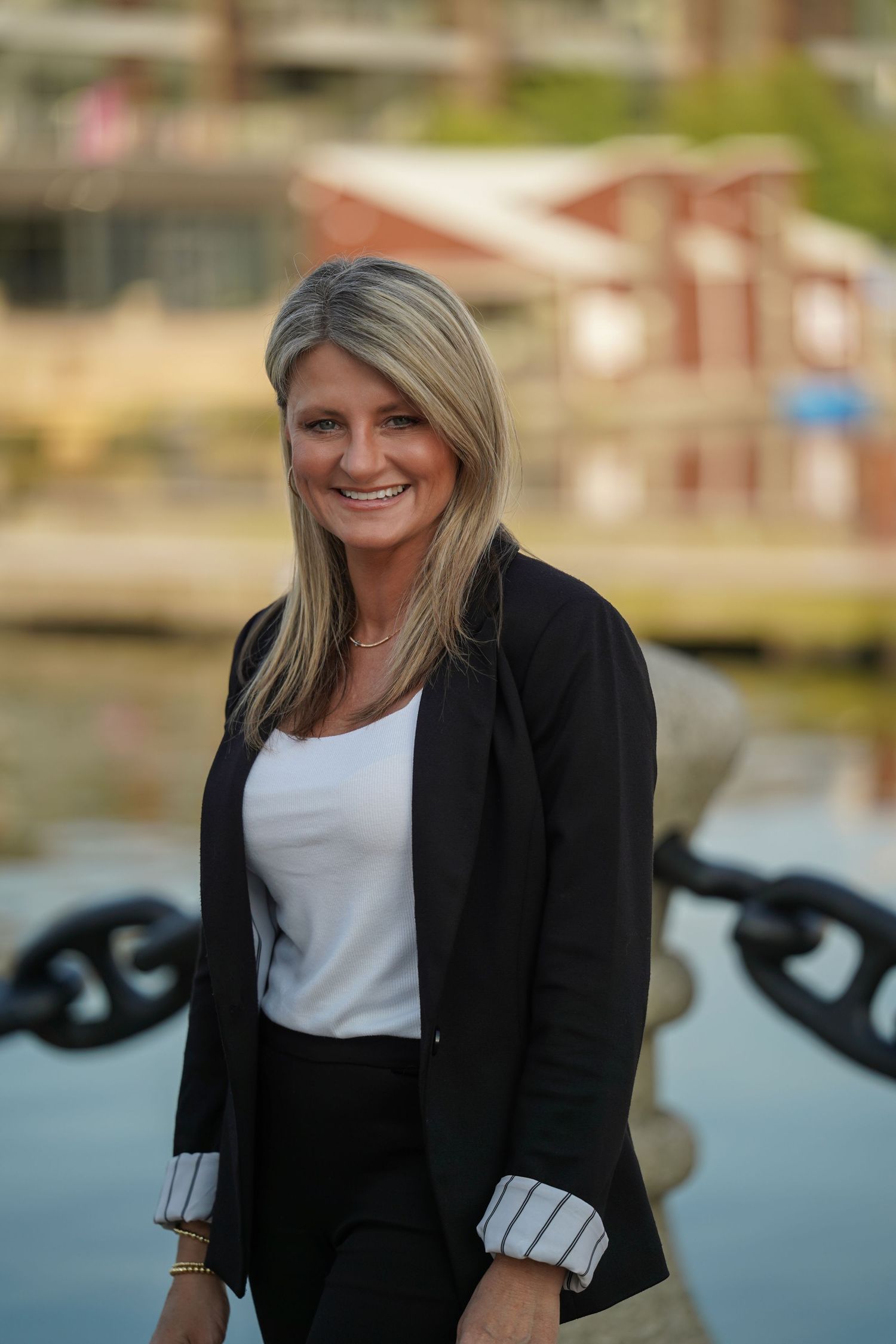 Professional portrait of a smiling woman in a black blazer and white top standing by a waterfront with boats in background.